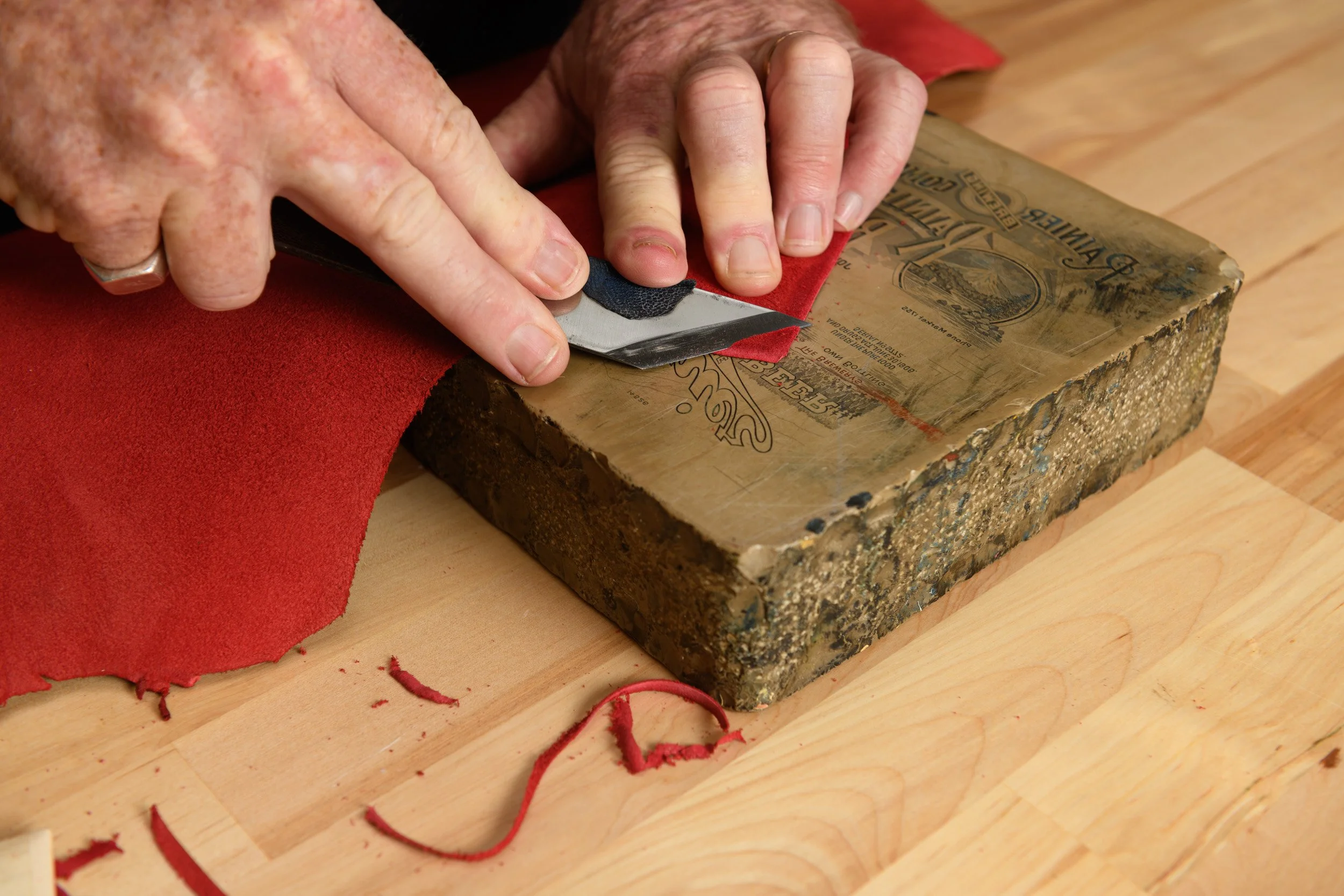 Close-up of hands using a utility knife to cut red material on a wooden block, with scraps of the material scattered around.