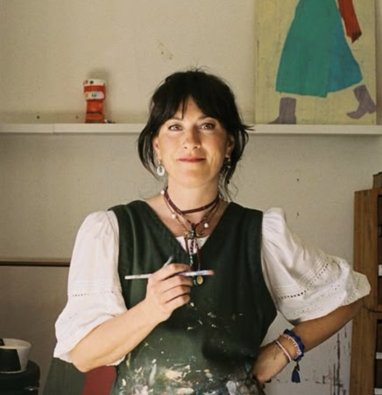 A woman with short dark hair, wearing a white blouse with puffed sleeves and a black apron, holds a paintbrush in her right hand. She is standing in an art studio with a shelf behind her that has a red figure and a painting of a person in a green skirt.