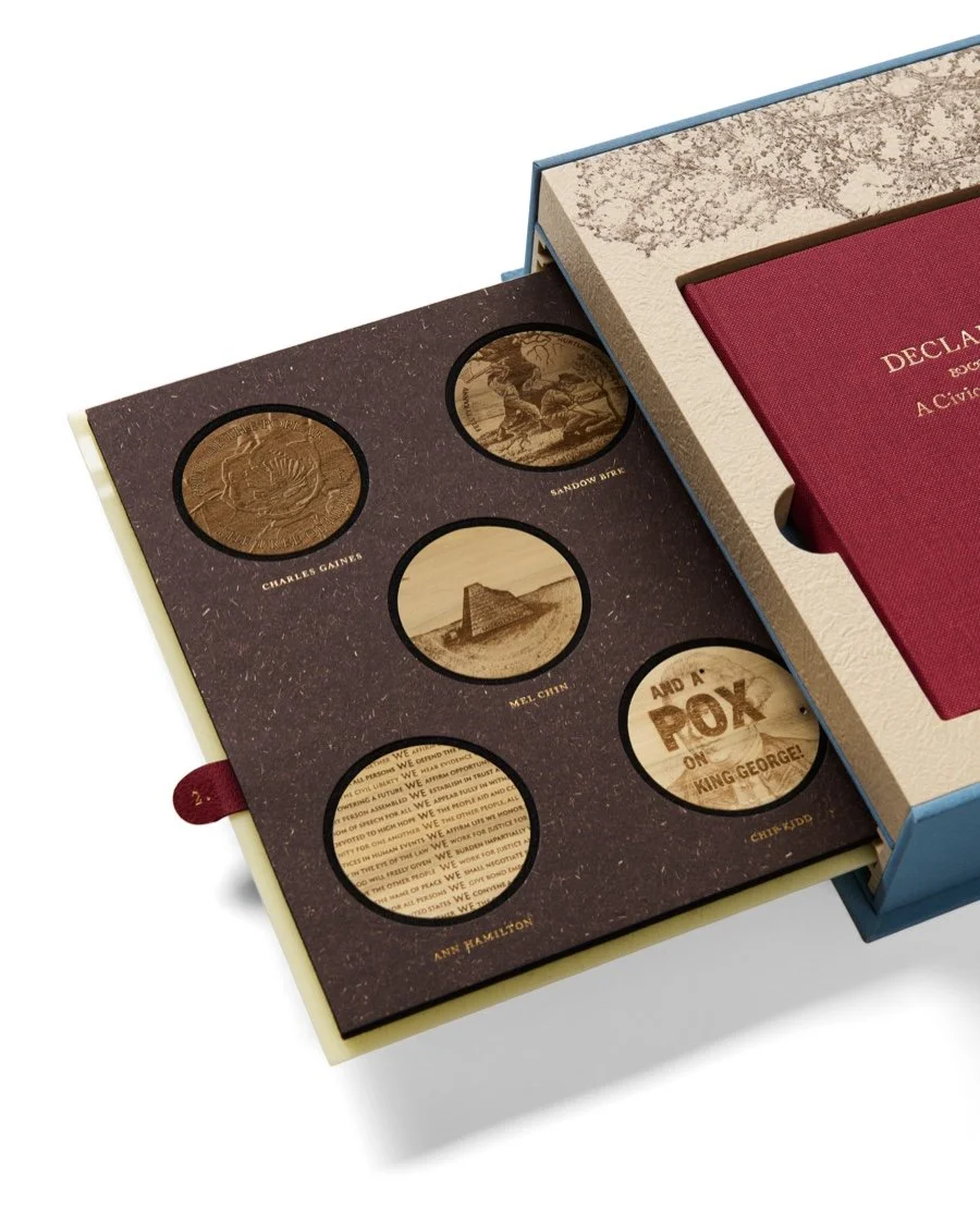 Open box with six circular cutouts displaying historical figures and scenes on wooden surfaces, related to American history, with a red book titled 'Declaration of Independence' inside the box.