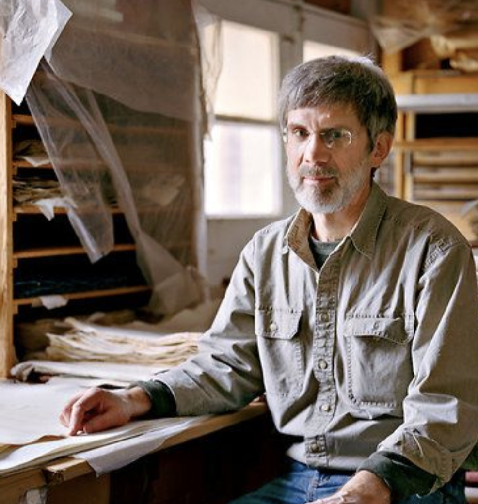 A middle-aged man with glasses and a beard sitting at a cluttered workspace with stacks of paper and wooden shelves in the background, in a workshop or studio.