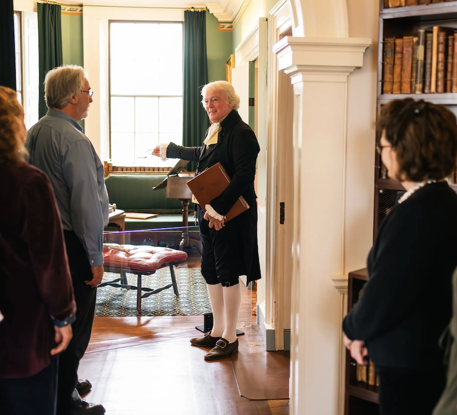 Group of people in historic room with large window, green curtains, and bookshelves, listening to a man dressed in 18th-century costume holding a folder.