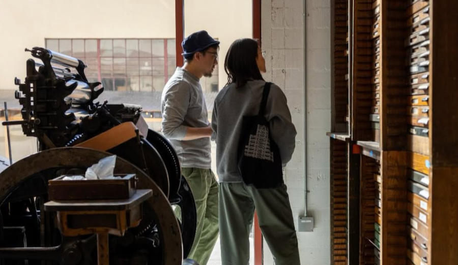 Two people standing by a wooden mail collection box, with the woman looking inside and the man looking on, in an indoor space with large windows.