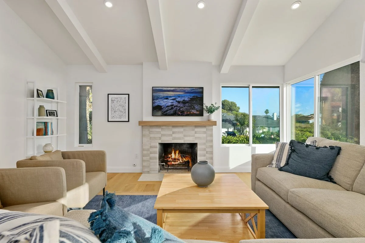Living room with a fireplace, a wall-mounted TV displaying a rocky shoreline, beige sofas with pillows, a wooden coffee table with a gray vase, large windows showing trees and a blue sky, and white walls.