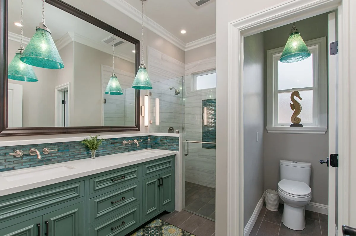 A bathroom with a double sink vanity, large mirror, and a walk-in shower with glass door. There is a small separate toilet room with a window and decorative seahorse statue.
