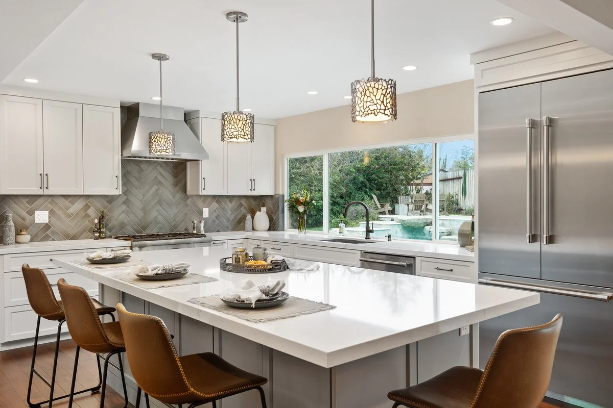 Modern kitchen with white cabinets, stainless steel refrigerator, a large island with place settings, pendant lights, and a view of a backyard with a pool.