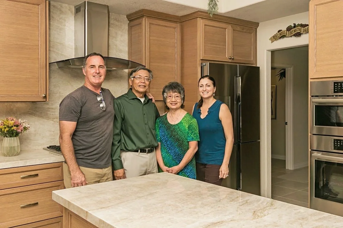 Four people standing together in a kitchen, smiling at the camera. The kitchen has wooden cabinets, a stainless steel refrigerator, and a beige countertop.