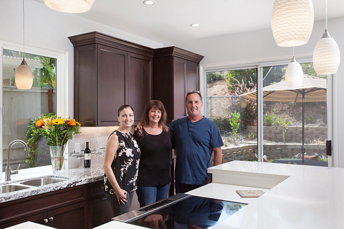 Three people standing in a modern kitchen with dark cabinets, white countertops, and large sliding glass doors leading outside, with a vase of colorful flowers and wine bottle on the counter.