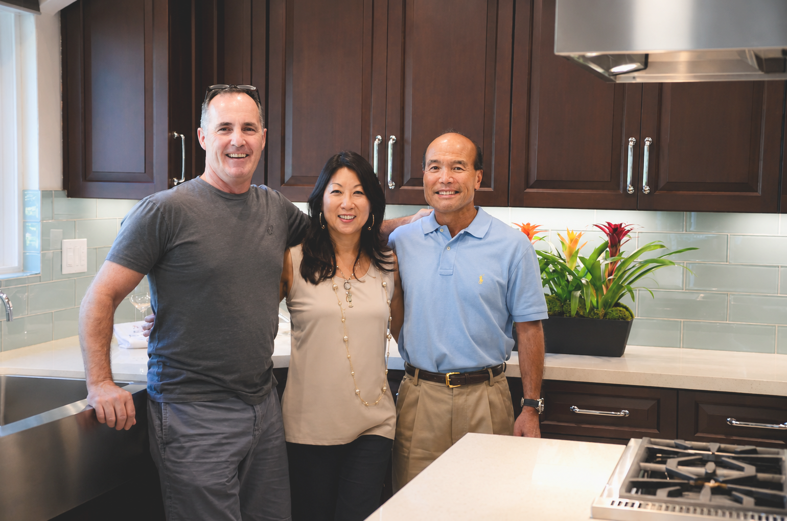 Three people standing in a kitchen, smiling at the camera. There are dark wood cabinets, a potted plant, and a stove in the background.