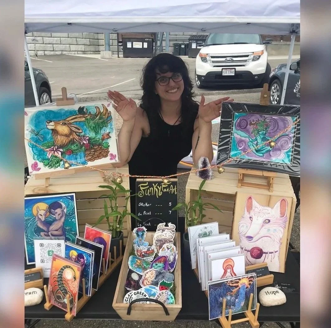 A person standing behind a booth displaying colorful artwork and stickers, with two framed art pieces featuring fantasy creatures, at an outdoor art fair.