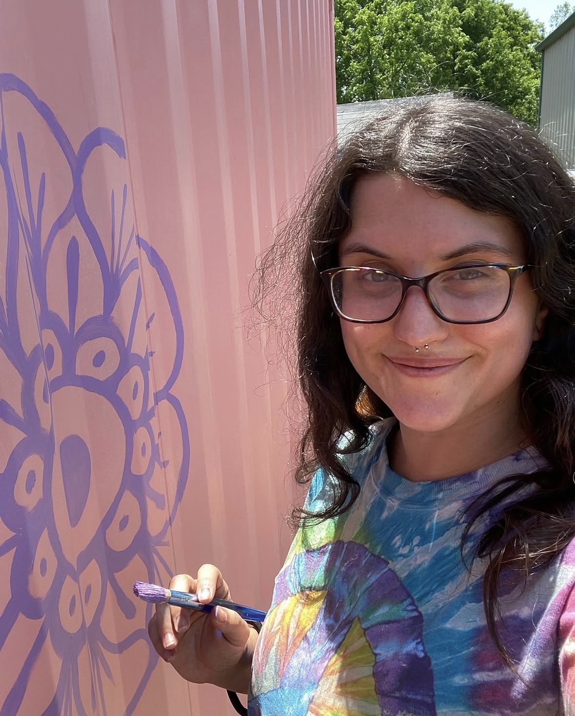 A young person with glasses and a septum piercing smiling while painting a purple flower design on a pink wall outside.