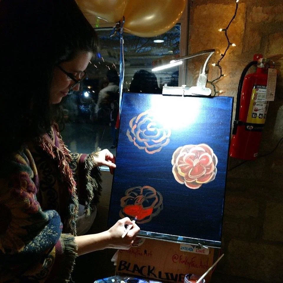 A person painting colorful roses on a dark background at a live event, with a fire extinguisher and string lights in the background.