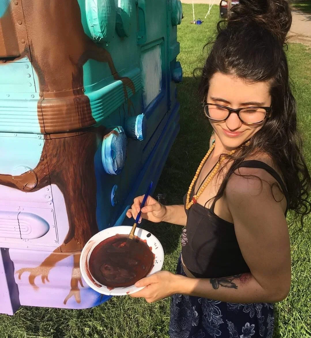 Young person with glasses and dark hair in a half bun painting a mural of a tree on a converted school bus, holding a palette with dark colors and a paintbrush.