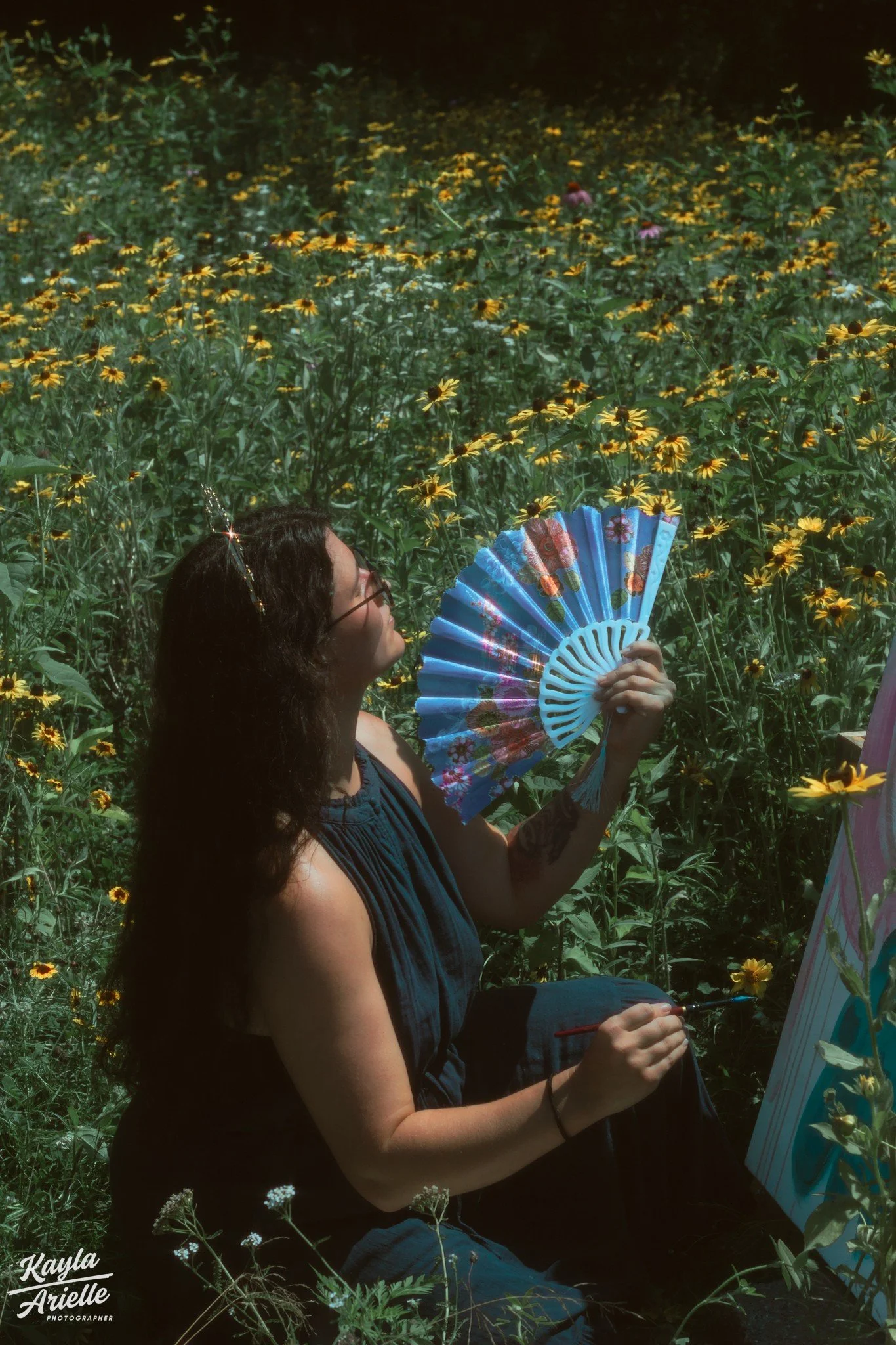 A person with dark curly hair, wearing glasses and a sleeveless dark blue dress, sits among yellow flowers, holding a colorful fan and a paintbrush, while working on an art piece outdoors.