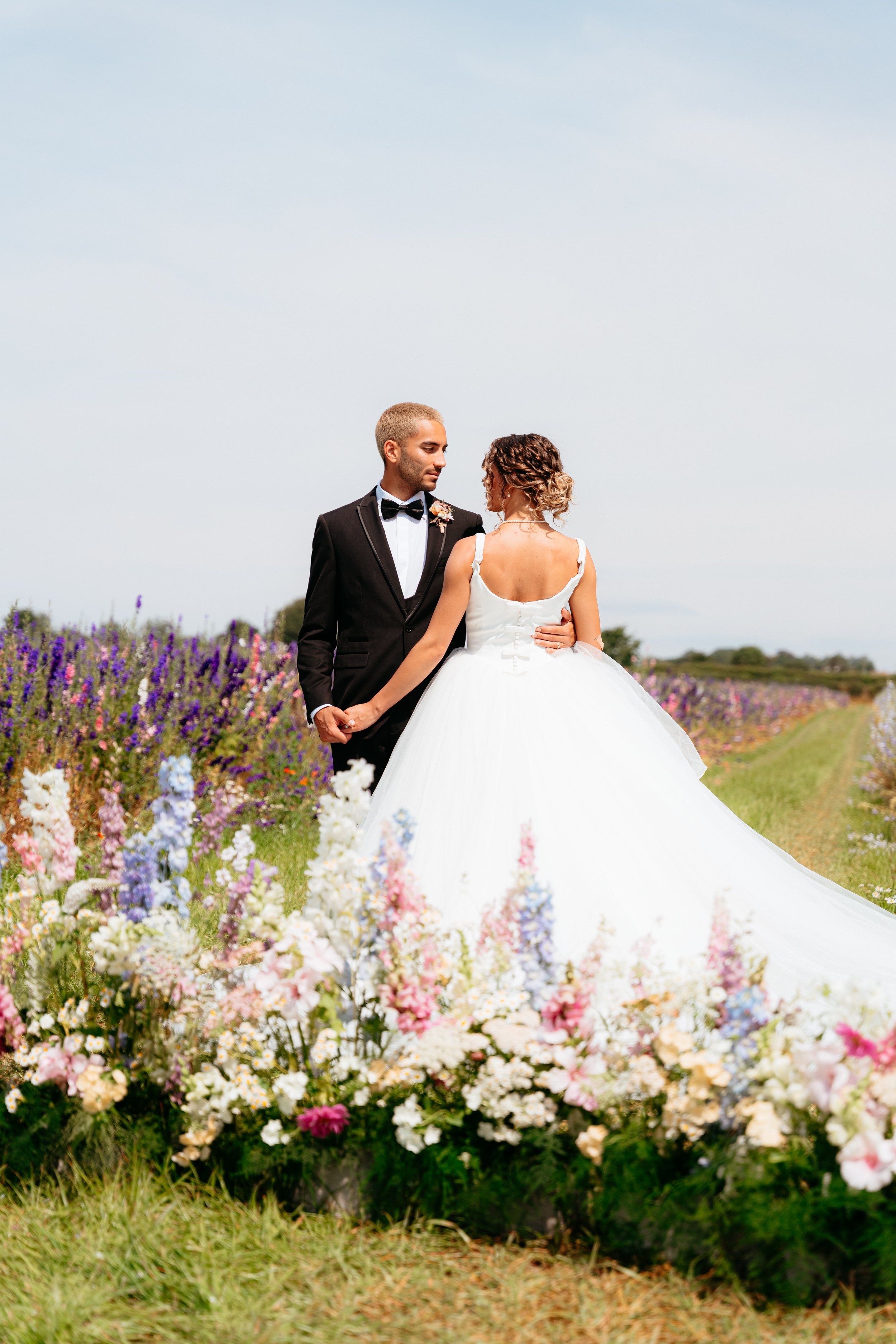 A bride and groom standing in a field of colorful flowers, holding hands and looking at each other on a sunny day. Gorgeous grounded floral arch as the ceremony backdrop in the beautiful Shropshire countryside.