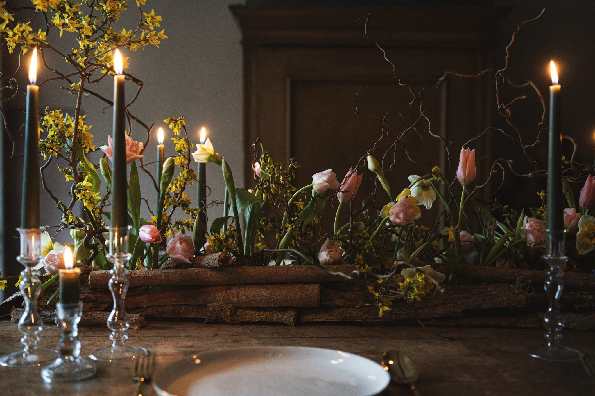 A wedding tablescape decorated with a rustic floral centerpiece of Tulips and Daffodils and yellow blossoms, surrounded by five tall, lit candles in glass holders. Perfect for a rustic Shropshire barn wedding.