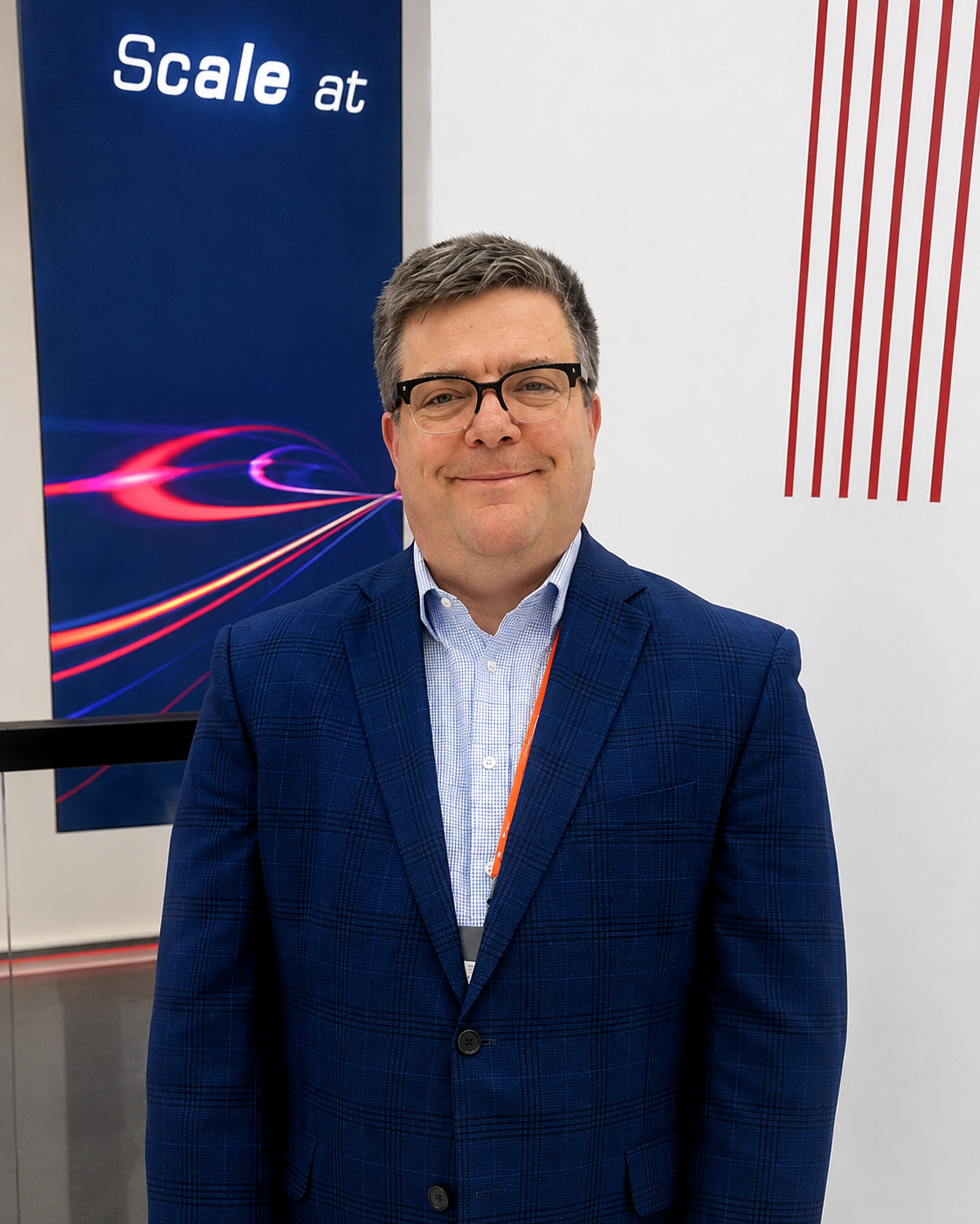 A man with glasses in a navy blue plaid suit and light blue shirt standing indoors in front of a blue sign that says 'Scale at' with colorful swoosh design, and red and white striped pattern on the wall to his right.
