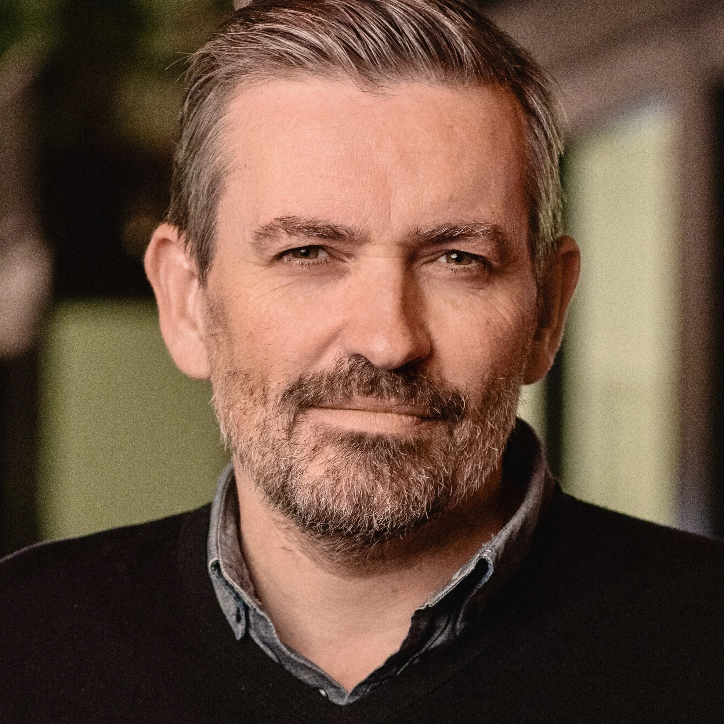 Close-up of a middle-aged man with gray hair and beard, wearing a black sweater and a collared shirt, with a blurred background.