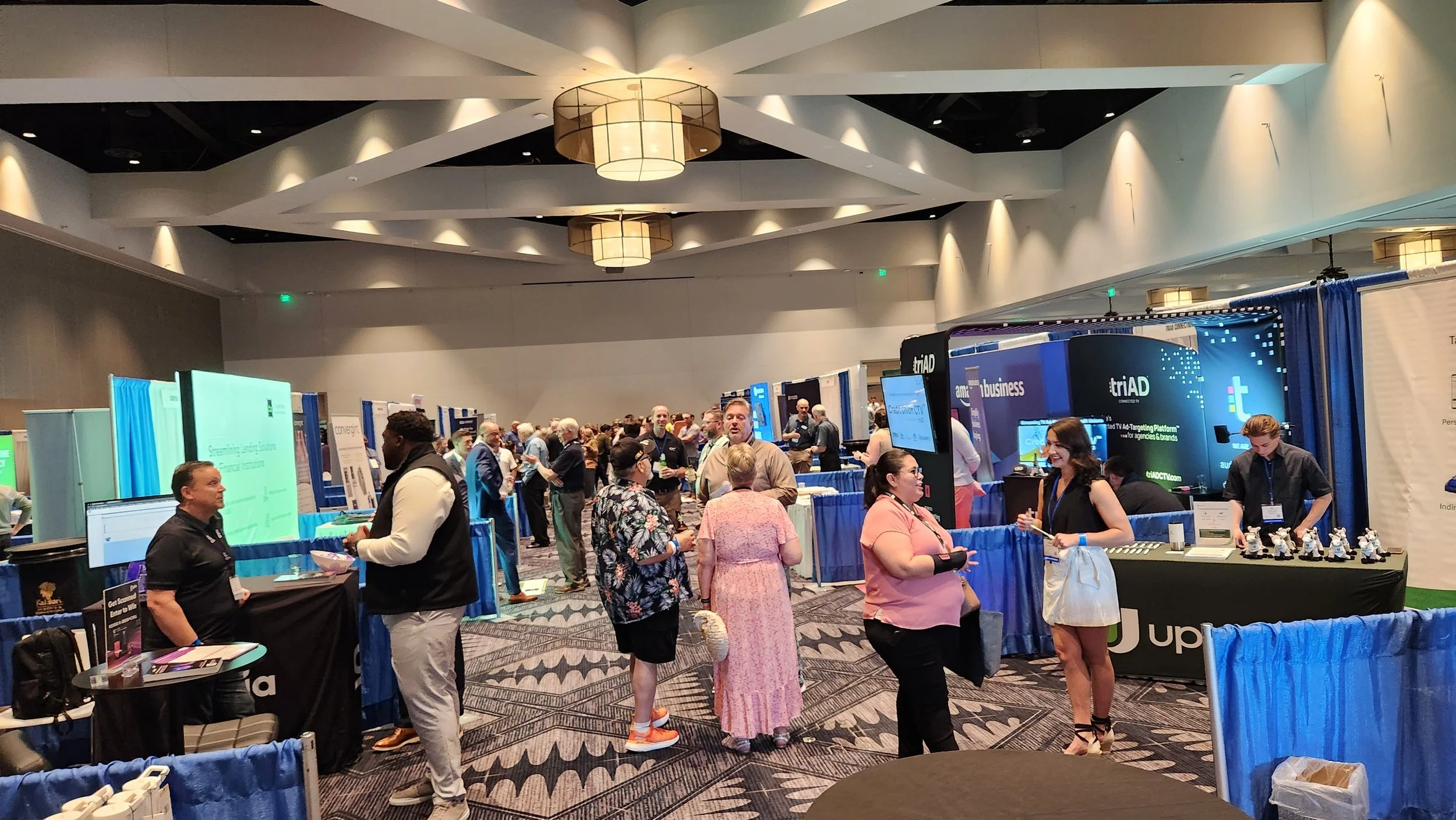An indoor conference or trade show with many booths and attendees. People are engaging with exhibitors and exploring displays, with a high ceiling and decorative lighting.