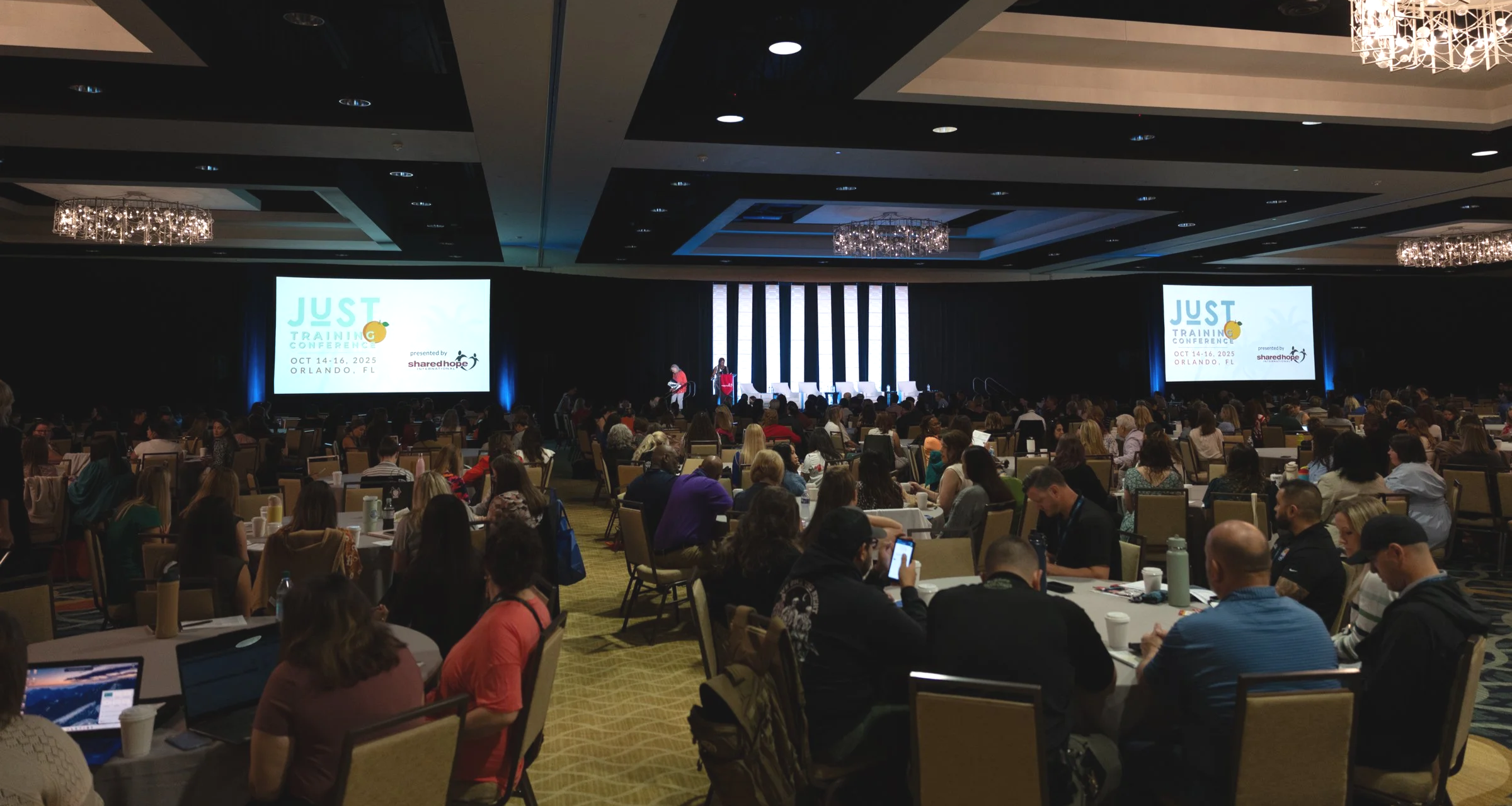 Conference room filled with seated attendees, large screens displaying the event name 'Just Training Conference' and details, stage with speakers, elegant ceiling chandeliers, and tables with laptops and water bottles.