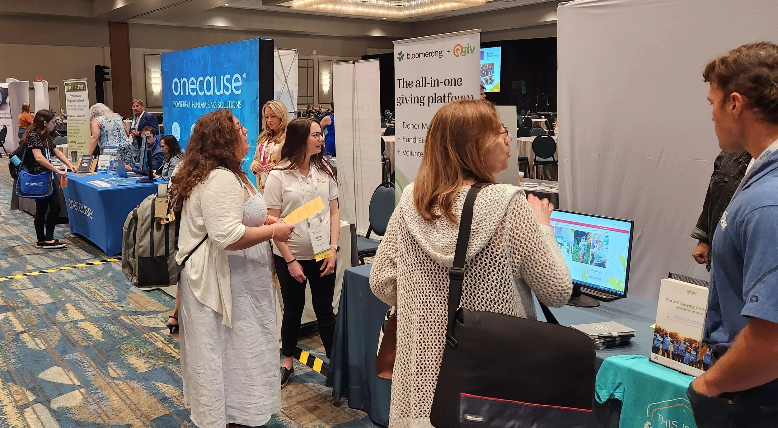 People attending a conference or event around a booth with banners and displays for various organizations, including 'onecause' and 'givr', in a large, well-lit meeting room.