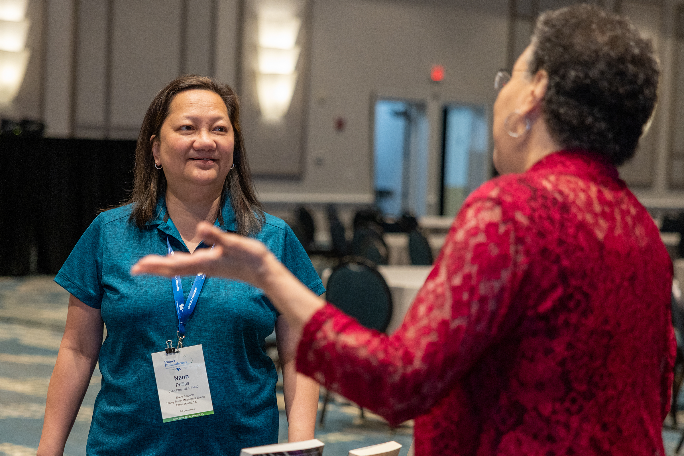 Two women engaging in conversation at a conference or event. One woman, in a blue shirt, is smiling and listening, while the other woman, in a red patterned shirt, is speaking with her hand raised. In the background, there are tables, chairs, and conference signage.