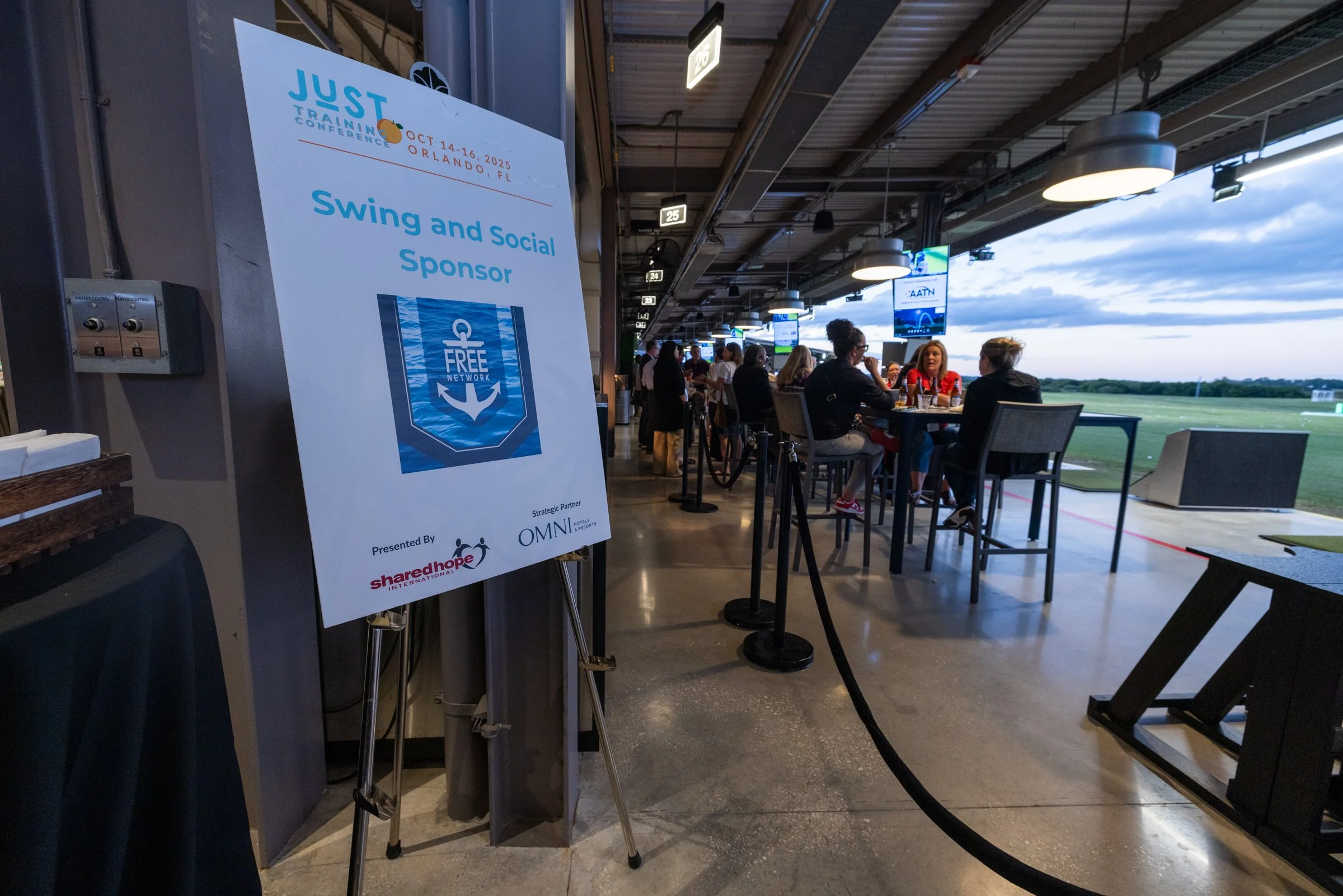 A sign at an indoor golf and social event sponsor area reading 'Swing and Social Sponsor' with logos including 'Share Hope International' and 'OMNI Hotels & Resorts.' The background shows people sitting at tables and watching golf swings outside, with screens displaying golf footage, under a ceiling with hanging lights.