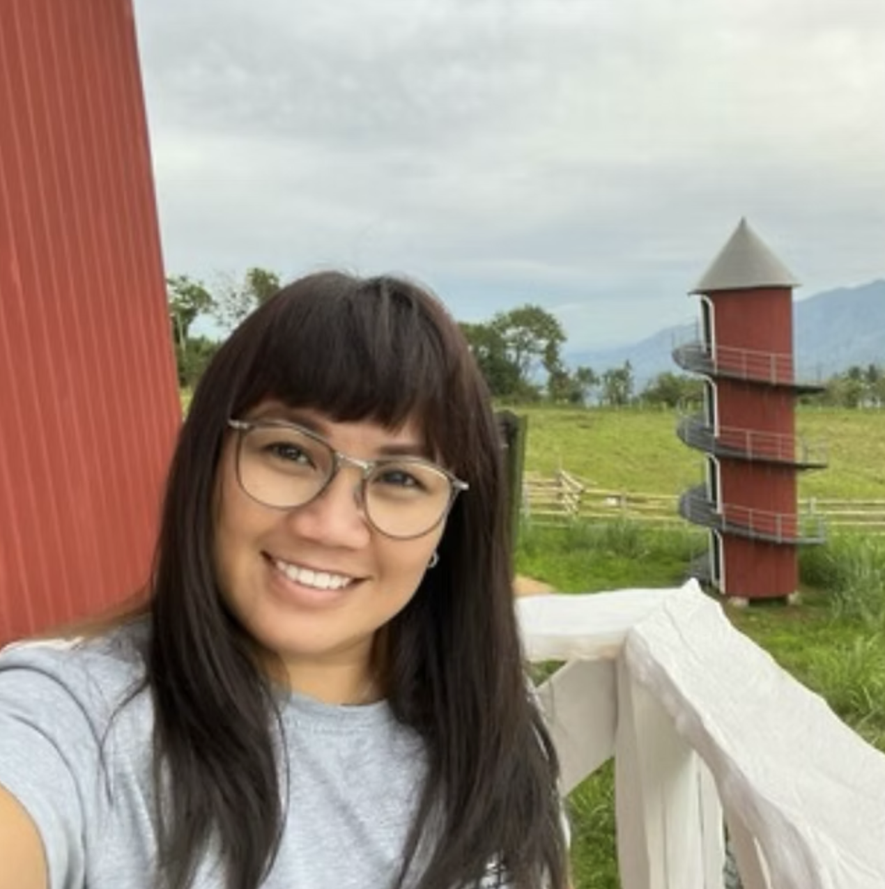 A woman with glasses smiling outdoors on a cloudy day, with a red spiral staircase and green landscape in the background.
