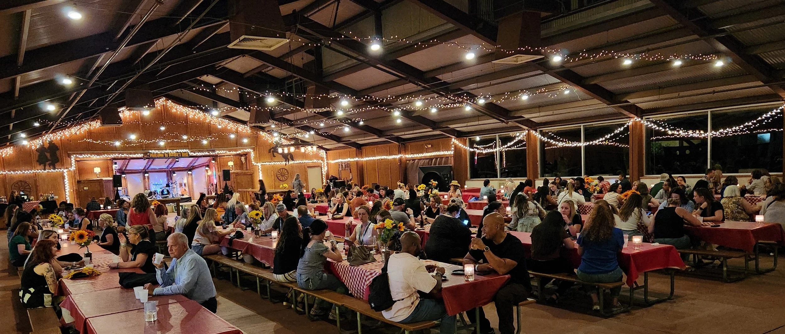 Indoor gathering with many people seated at long tables covered with red checkered tablecloths, decorated with sunflower centerpieces, under a ceiling with string lights. There's stage with musical instruments and a sign that reads 'Welcome to Rutter