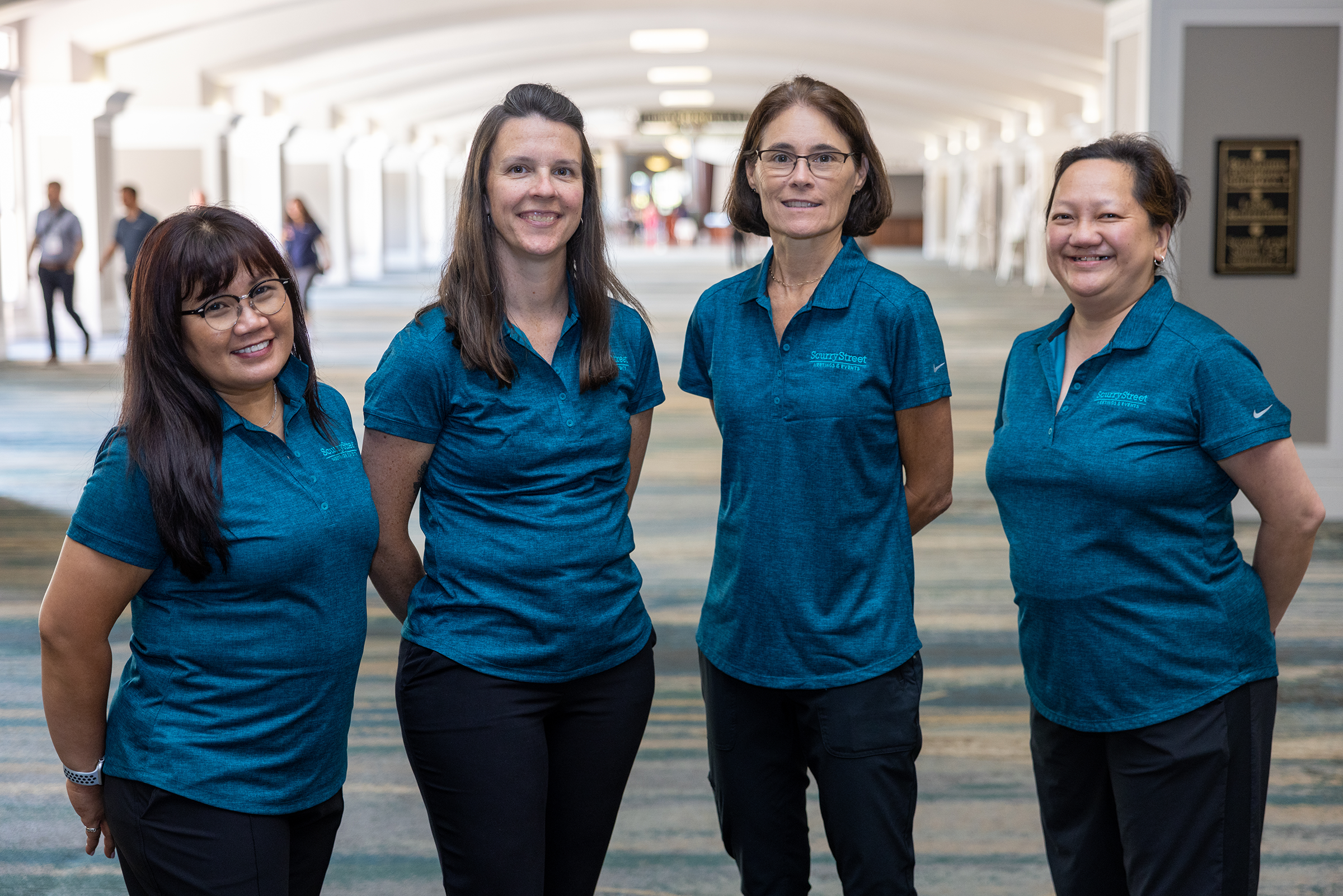 Four women wearing blue shirts standing together in an indoor hallway, smiling at the camera.