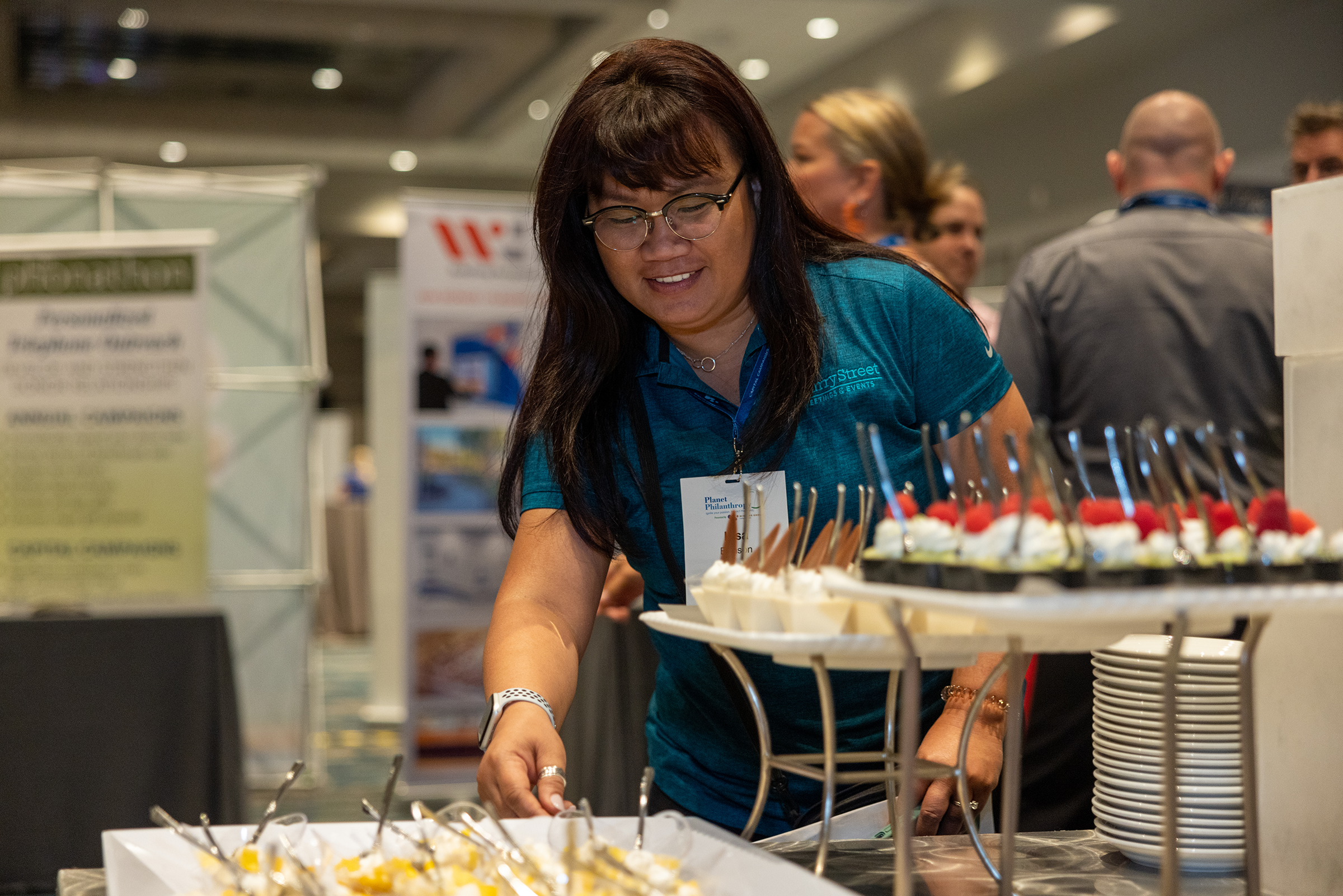 A woman with dark hair and glasses serving herself at a dessert buffet, with plates of cheesecake and cherry-topped desserts on display.