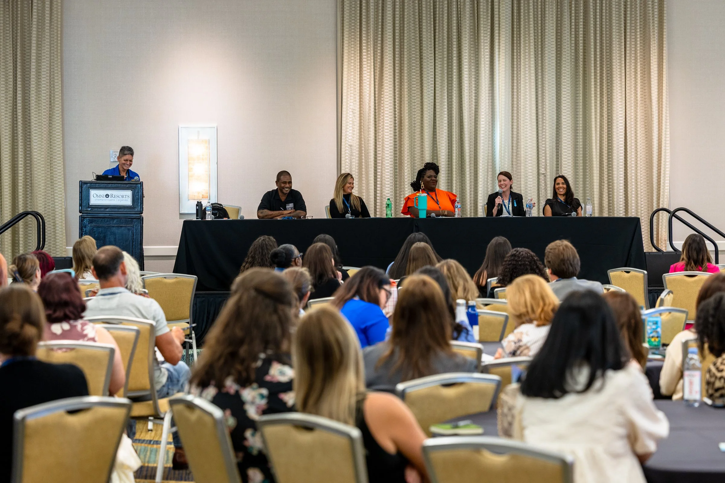 A panel of six people sitting at a table on a stage, with a woman standing at a podium on the left, in front of an audience in a conference room.