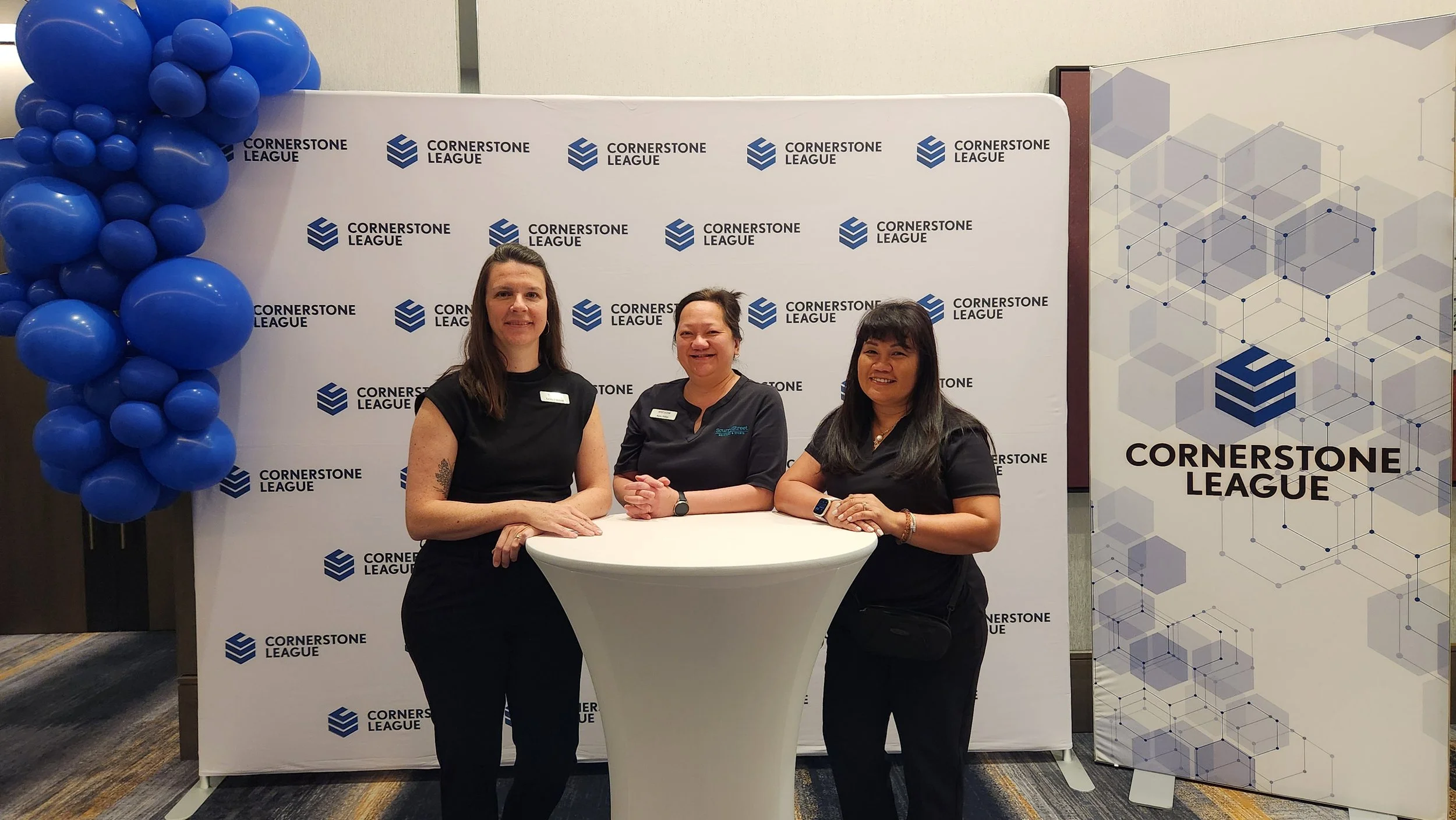 Three women standing behind a round table at a Cornerstone League event, with a backdrop and blue balloon decoration to the left.