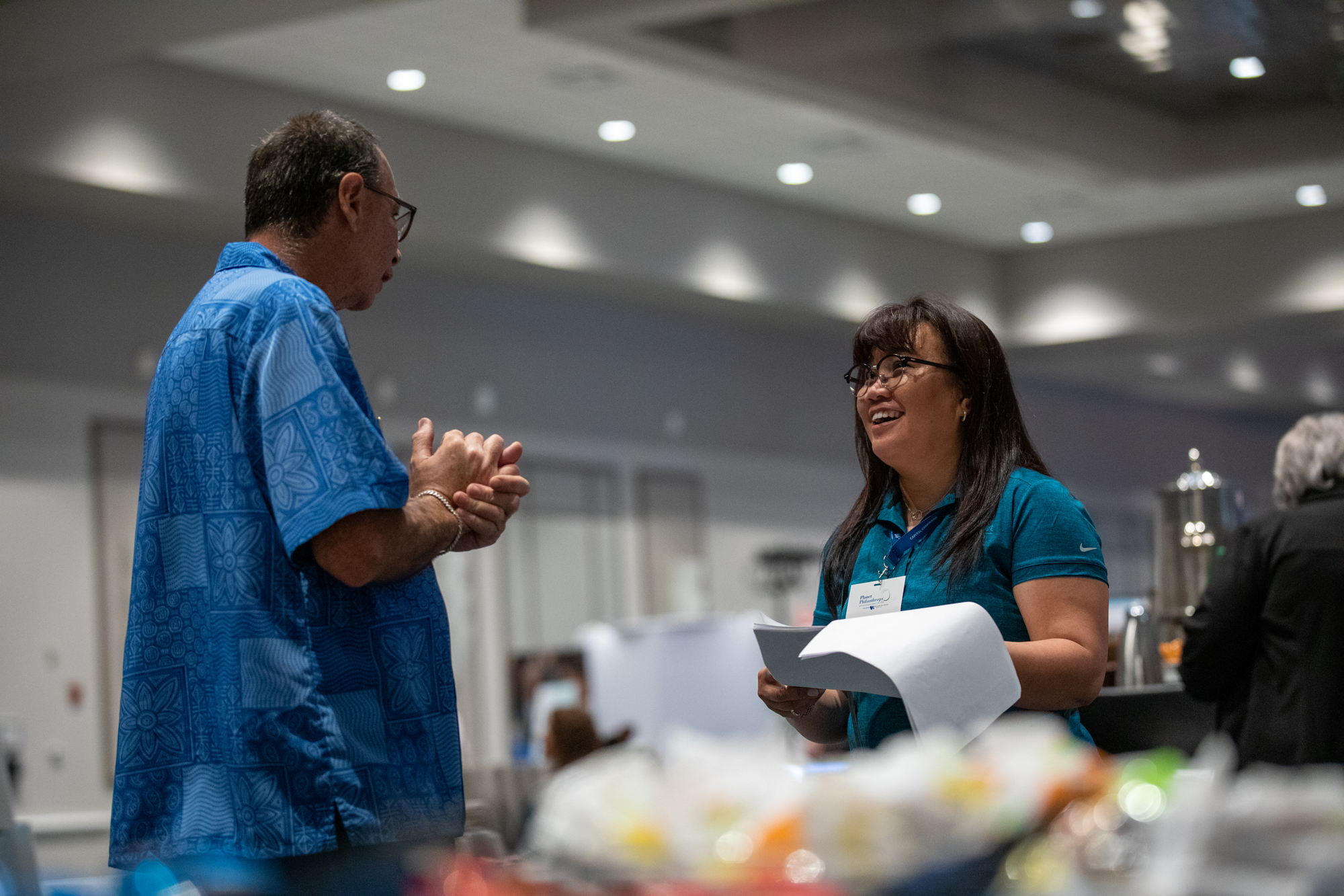 A man and a woman are engaged in conversation at an indoor event. The man is wearing glasses and a blue patterned shirt, and the woman is wearing glasses and a blue shirt. They are smiling and holding papers, with a blurred background of other people and refreshments.