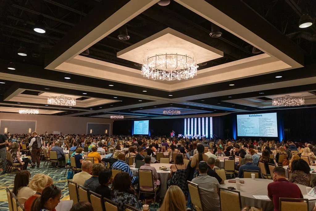 A large conference room filled with many attendees sitting at round tables, facing a stage with a large screen displaying a presentation. The room has ornate chandeliers and a high ceiling.