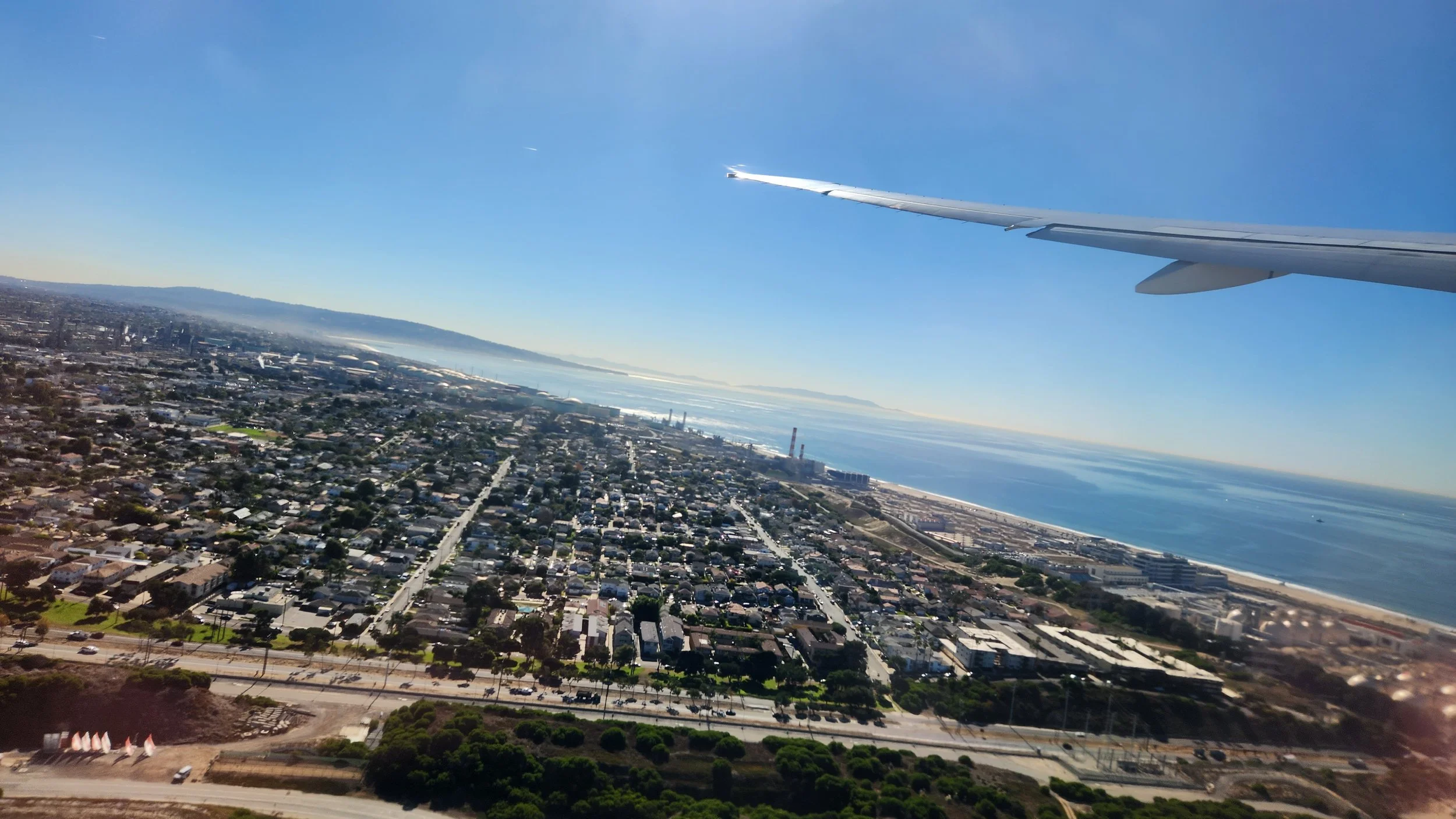 View from an airplane window showing urban area and coastline below, with airplane wing visible in the upper right corner.