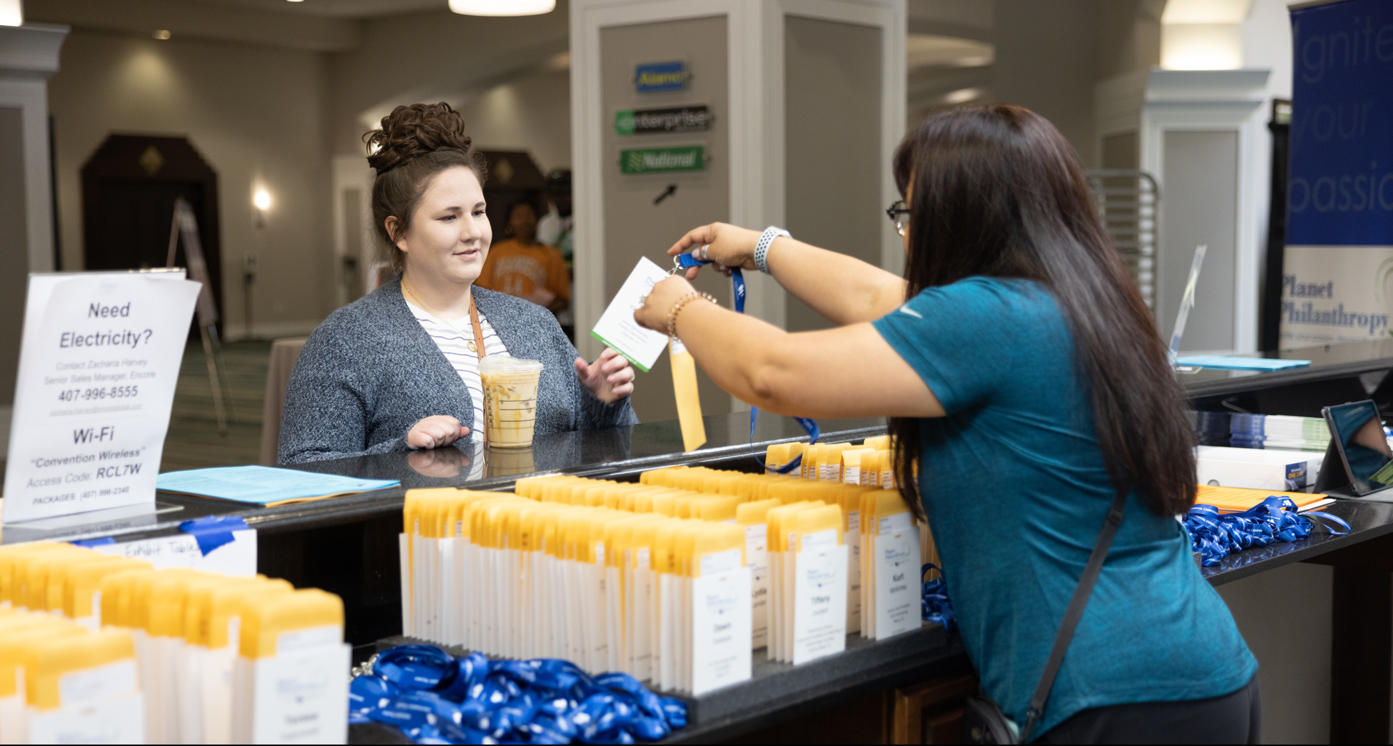 A woman with glasses and a blue shirt hands a sticker or badge to a young woman with curly hair styled in an updo, wearing a striped shirt and a gray sweater, who holds a drink. They are at a registration table with yellow badges and blue lanyards, and there are informational signs about electricity and Wi-Fi on the table.