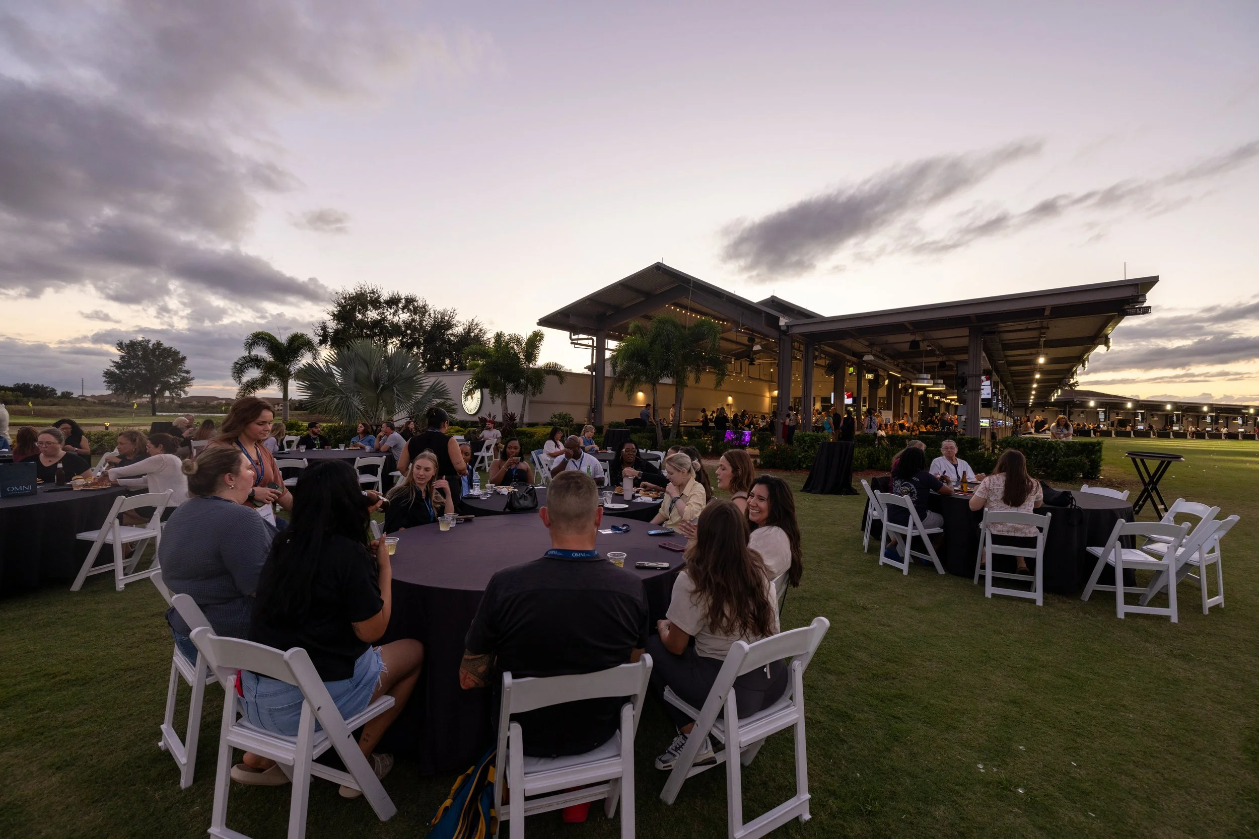 Outdoor event with people sitting at round tables on grass, some eating, others talking, under a modern structure with palm trees in the background, during sunset with cloudy sky.