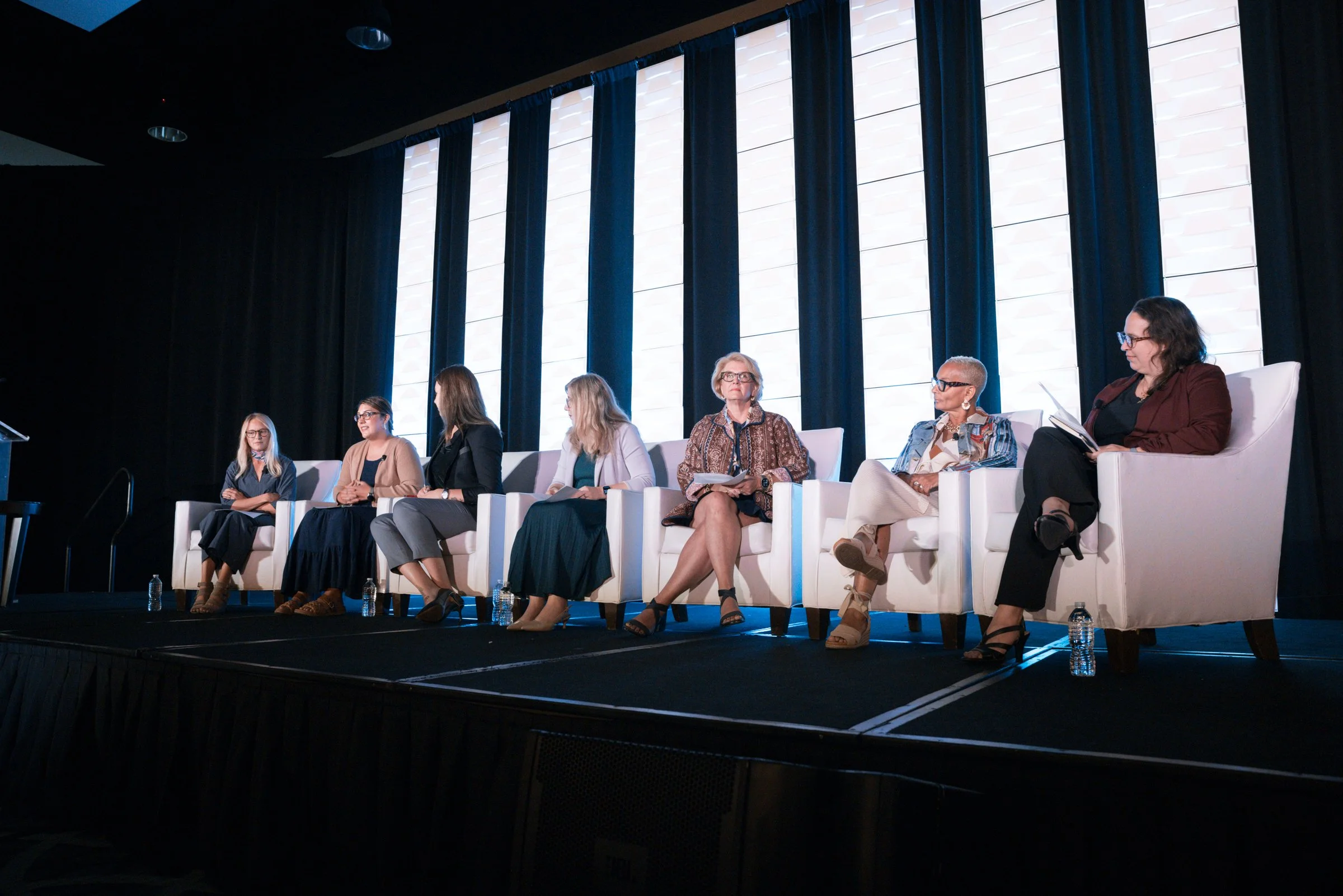 Eight women sitting on a stage in front of blue curtains, participating in a panel discussion, with water bottles at their feet, in a large auditorium.