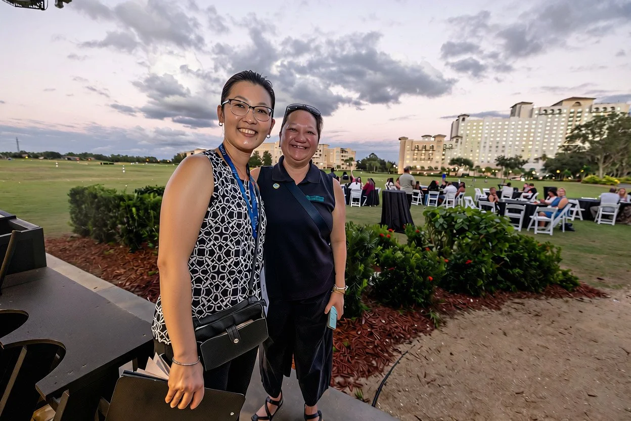 Two women smiling outdoors at a social event during sunset, with tables and chairs in the background on a grassy field surrounded by bushes and large buildings.