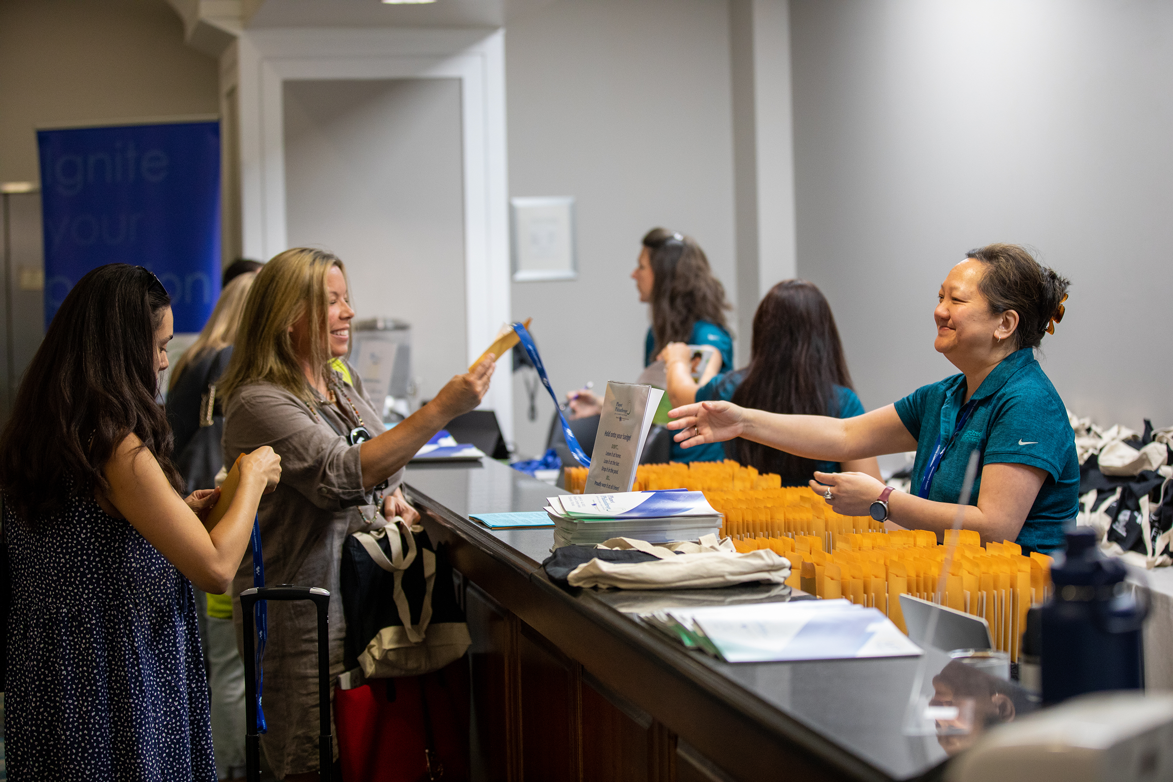 Reception area with women attending an event, handing or receiving yellow envelopes at a registration table with brochures and lanyards, in a well-lit room.