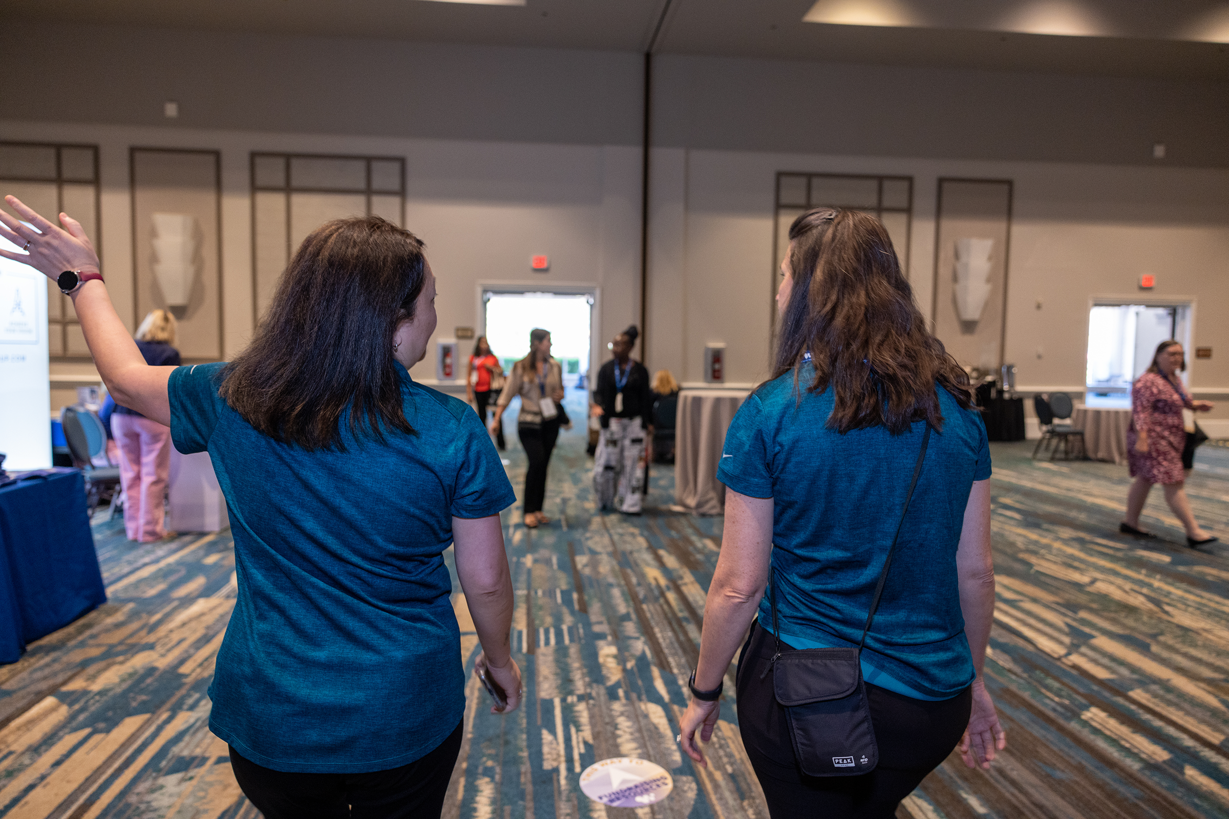Two women walking through an indoor conference or event with booths and other attendees in the background.