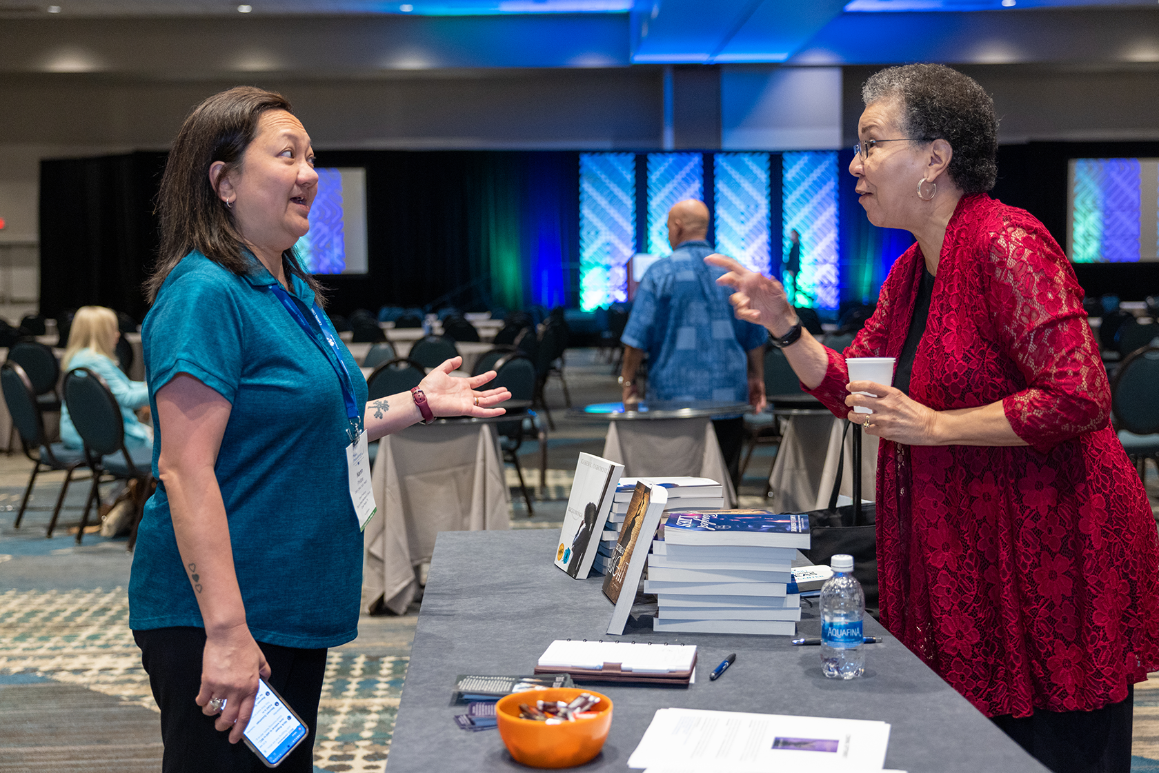 Two women are having a conversation at a book signing event in a conference hall. The woman on the left is wearing a blue polo shirt and holding a mobile phone, while the woman on the right is wearing a red lace jacket and holding a cup. There are books on a table between them and other attendees in the background.