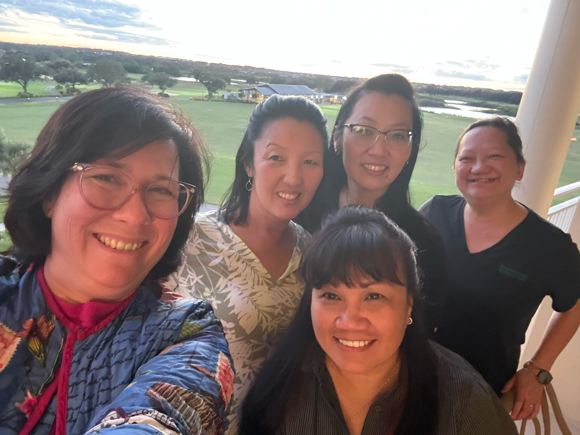 Group of five women smiling and taking a selfie outside near a golf course at sunset.