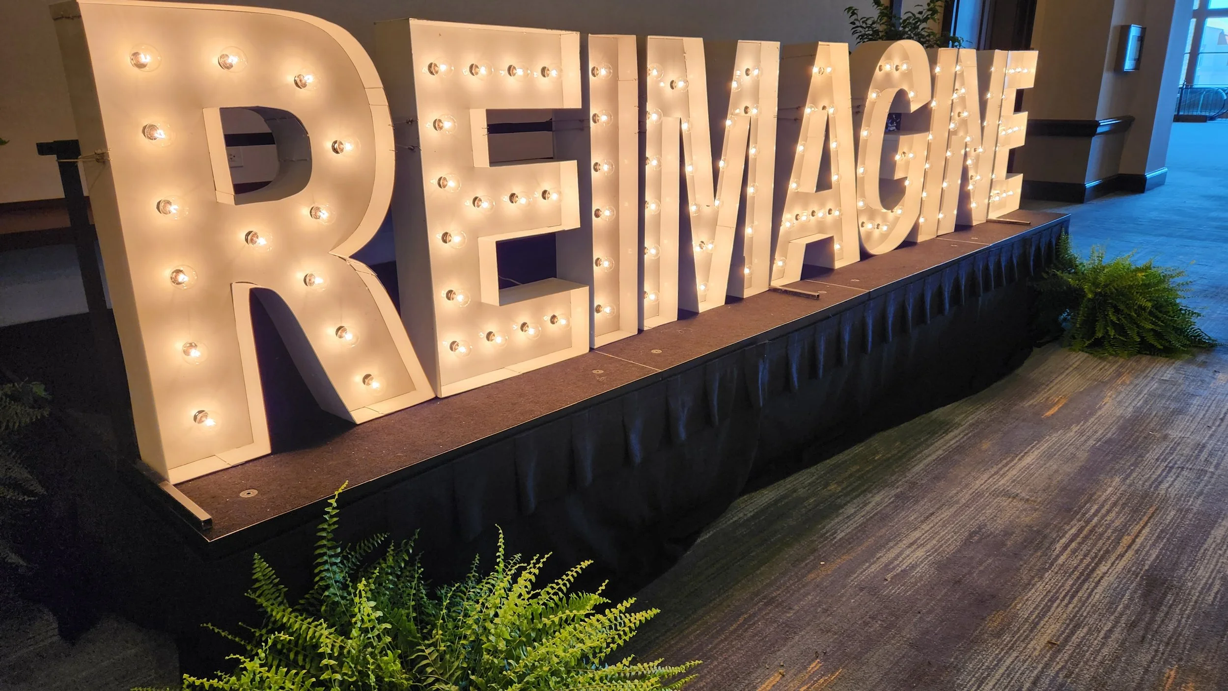 Large marquee sign spelling out 'REVENGE' with white background and illuminated bulbs, situated on a raised platform indoors, with some greenery in the surroundings.