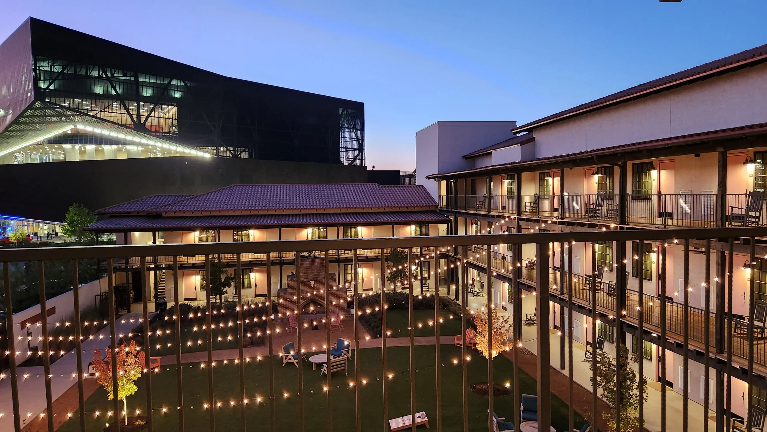 A courtyard view of apartment buildings with string lights, patio furniture, and a small lawn, with a modern glass building in the background during twilight.
