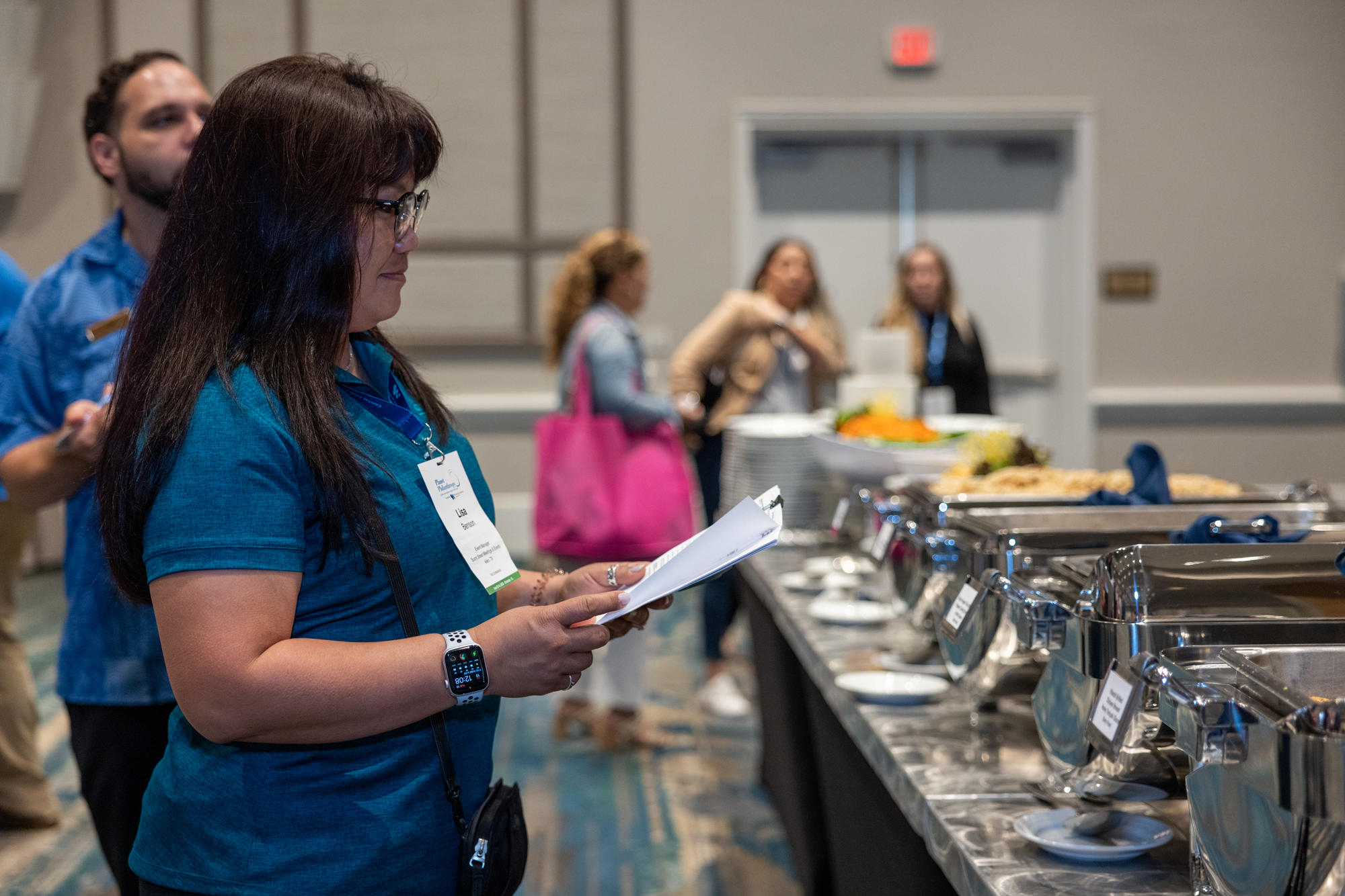 Woman with glasses and a wristwatch reading a snack brochure at a buffet table.