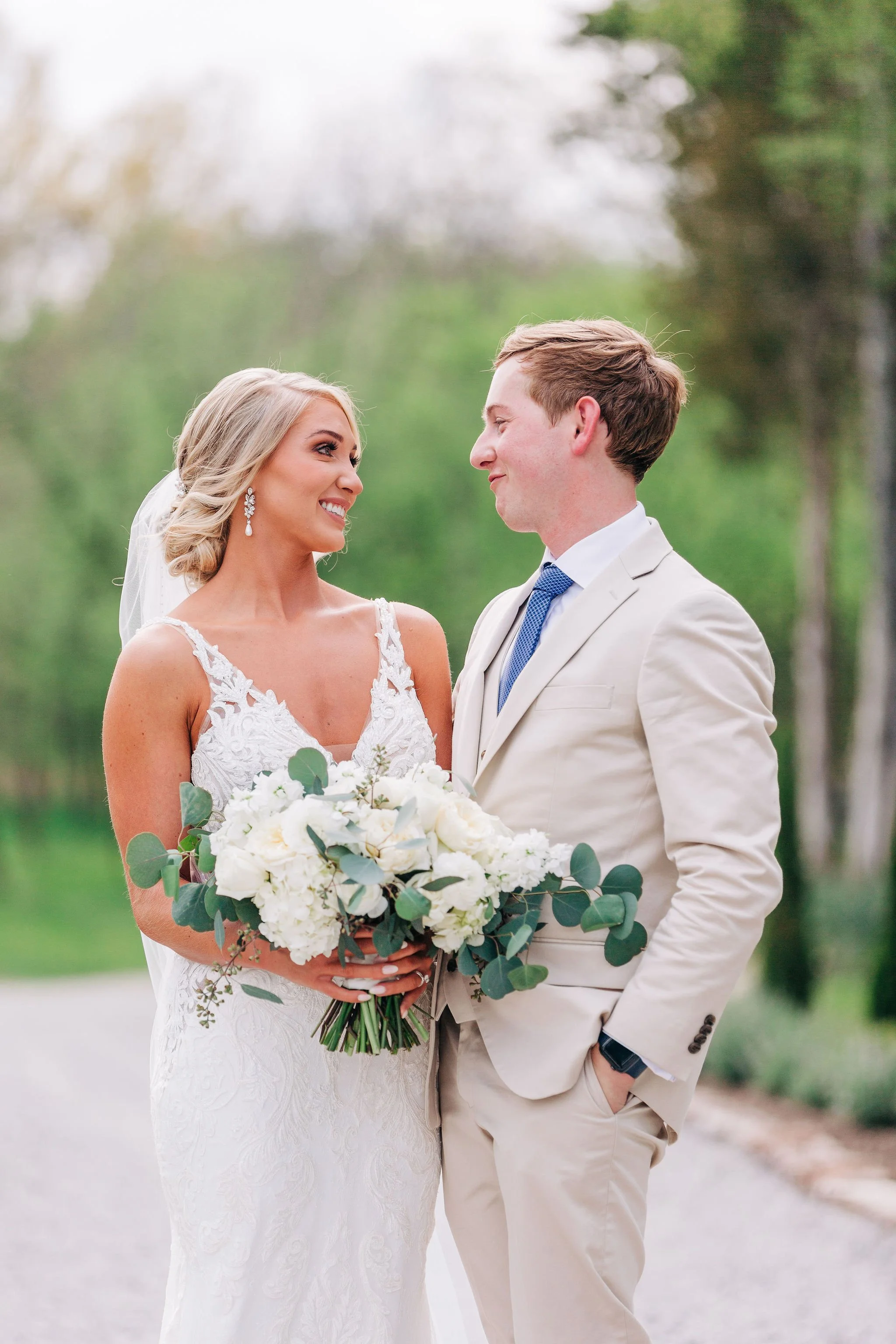 A bride and groom outdoors on a wedding day, smiling at each other. The bride is holding a large bouquet of white flowers and greenery, wearing a lace wedding dress and teardrop earrings. The groom is dressed in a light-colored suit with a blue tie and has his hand in his pocket, standing close to the bride.