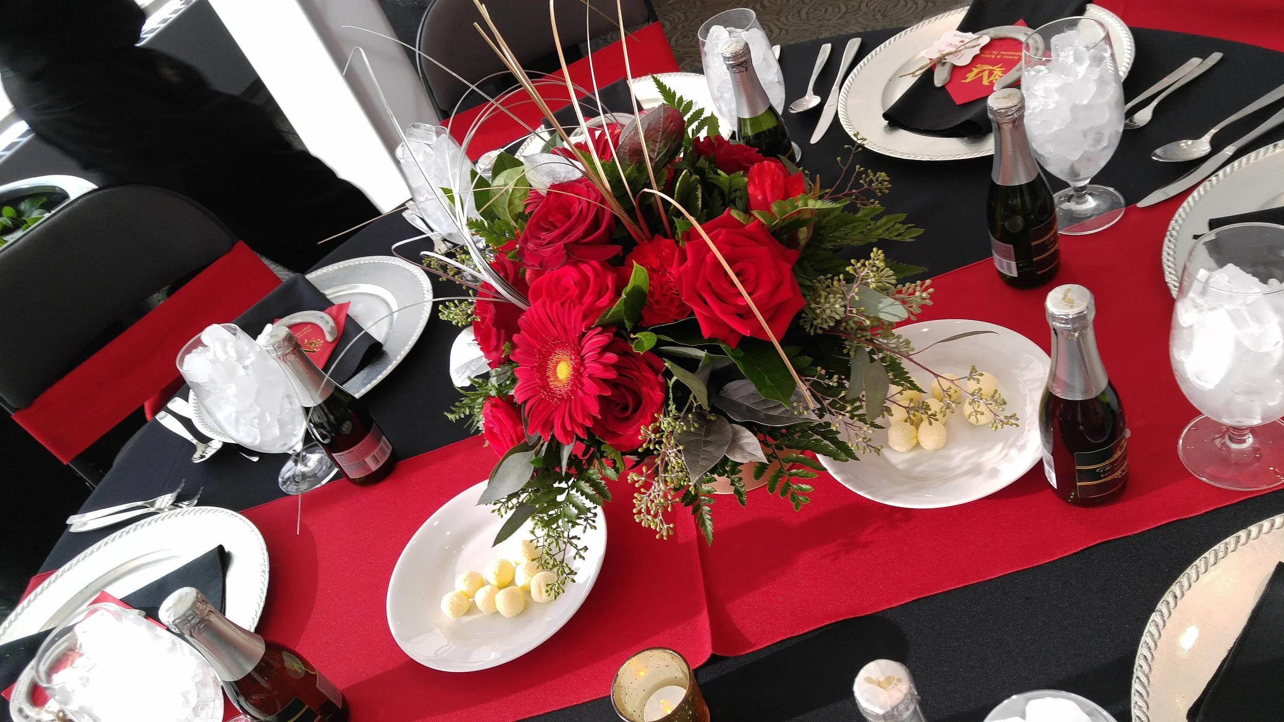 A table decorated for a celebration with a centerpiece of red roses, pink gerbera daisies, and greenery. The table is set with white plates, silverware, glasses filled with ice, and small bottles of sparkling cider or soda. There are also white bowls with what appears to be small round candies. The table has a black tablecloth with a red table runner, and the place settings include black and red napkins with gold accents.