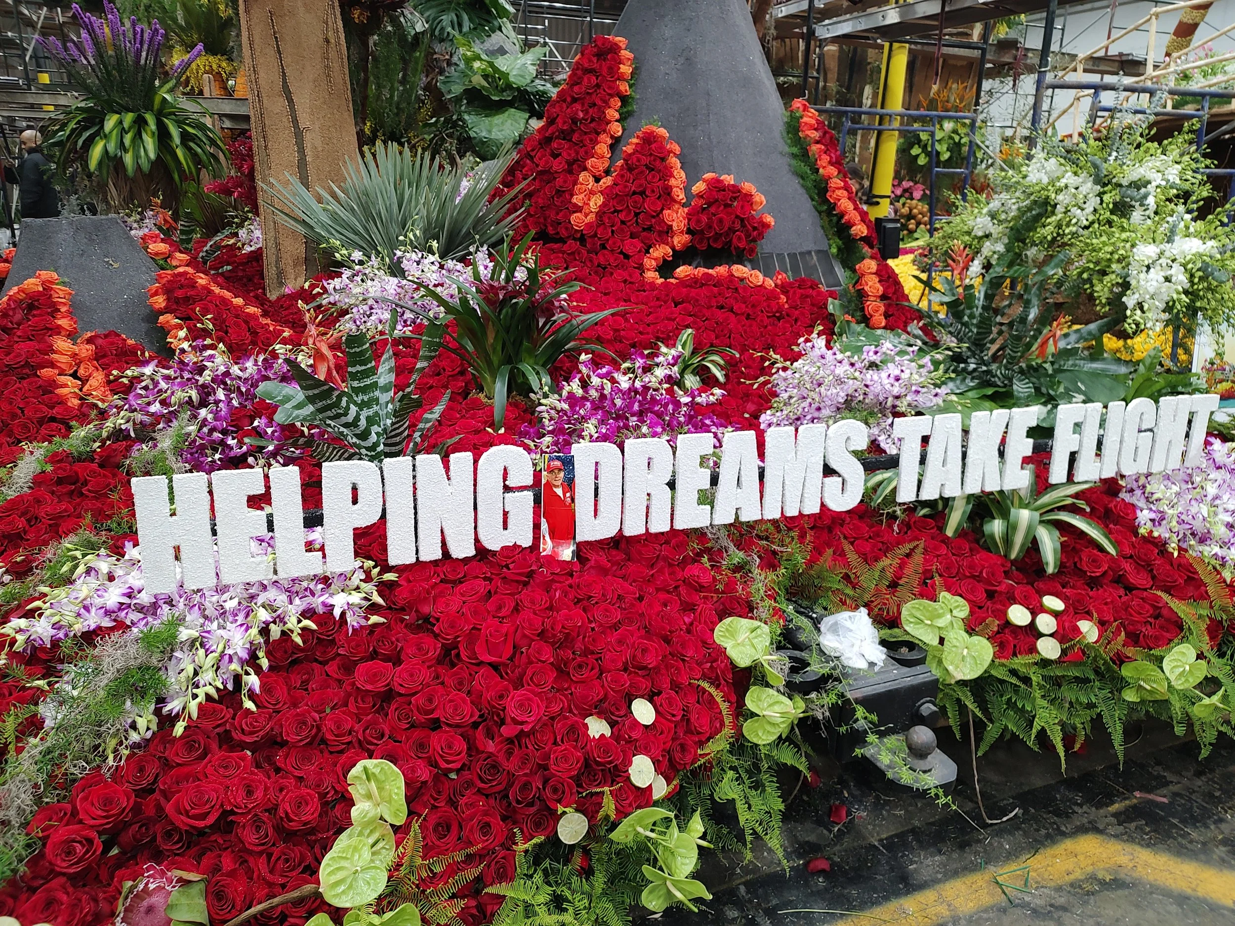 A display of various flowers, primarily red roses, with some orange and purple flowers, green foliage, and a sign with large white letters partially visible that reads 'HELP RANGERS DREAM'.