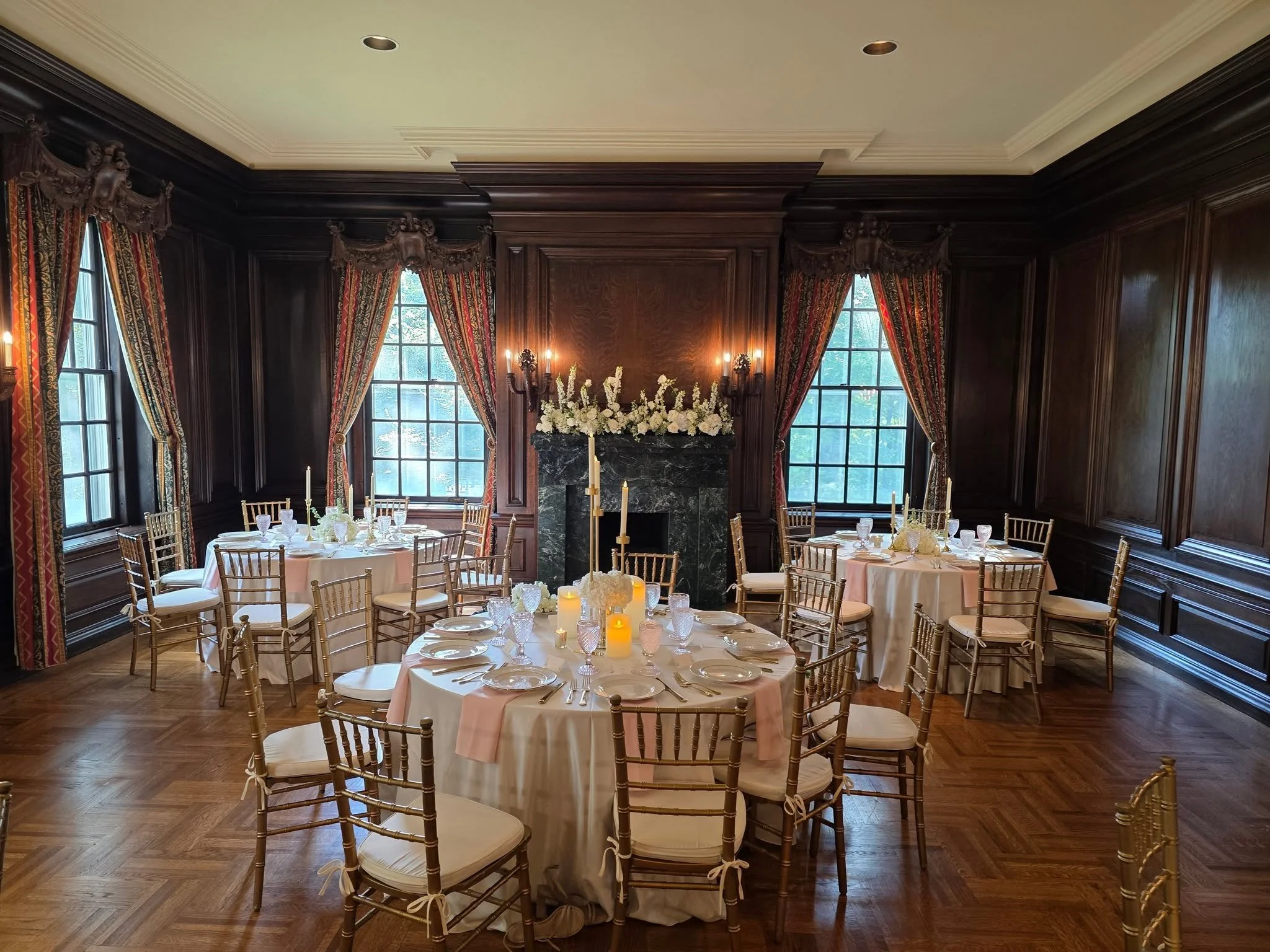 Elegant banquet room with round tables covered in white and pink tablecloths, decorated with candles and floral centerpieces, surrounded by wooden chairs, dark wood paneled walls, and large windows draped with ornate red and gold curtains, with a fireplace and floral arrangement in the background.